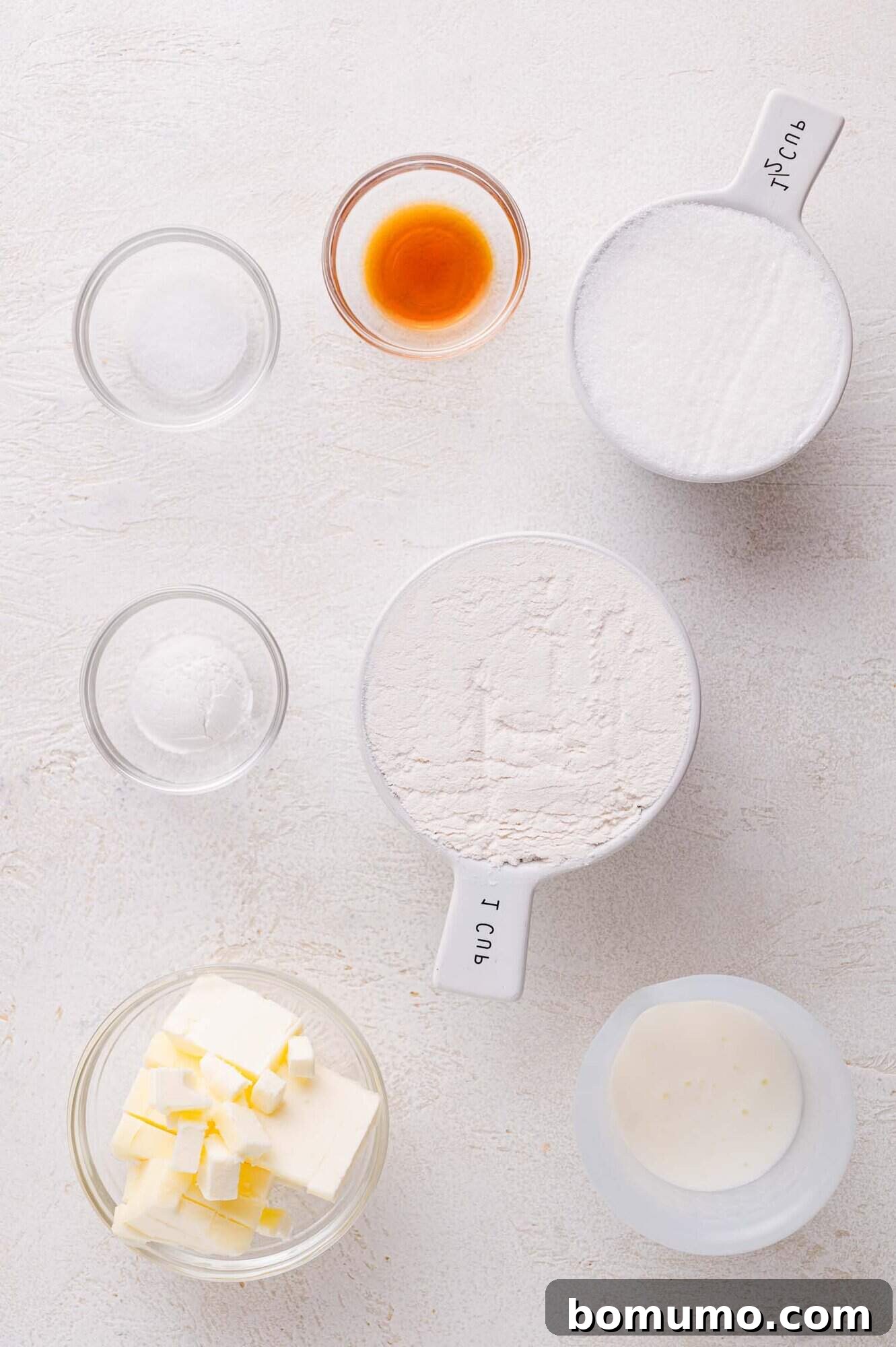 A collection of essential baking ingredients for the cobbler topping: flour, granulated sugar, cold butter cubes, rich buttermilk, aromatic vanilla extract, baking powder, and salt, artfully arranged in bowls on a clean white backdrop.