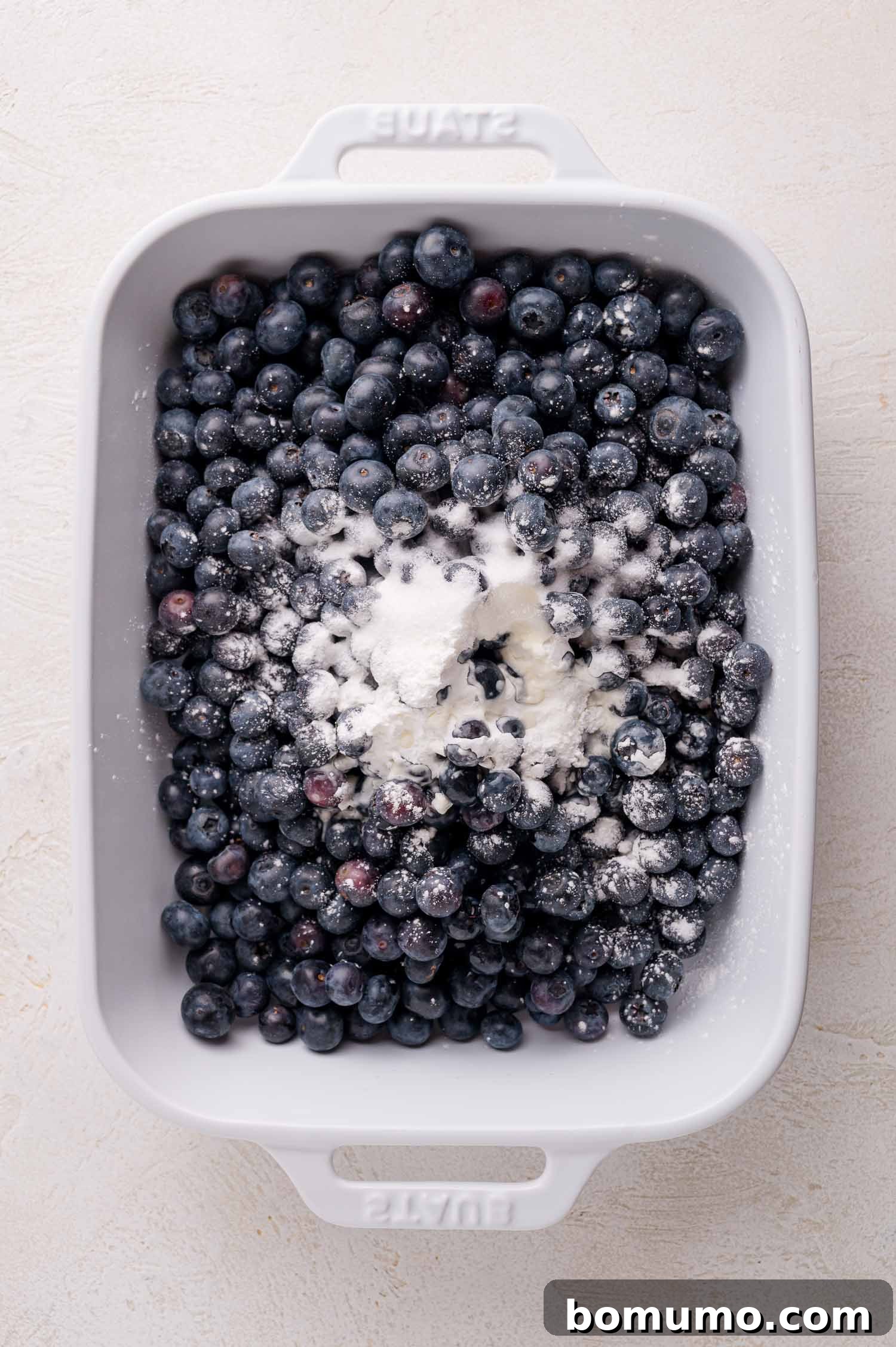 A white rectangular baking dish filled with a vibrant base of fresh blueberries, generously topped with a mixture of sugar and cornstarch, ready for preparation.