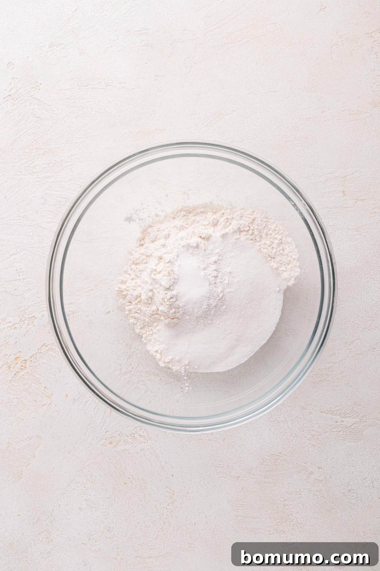 A clear glass bowl showcasing a harmonious mound of white all-purpose flour and a delicate sprinkle of granulated sugar, set against a subtly textured, light-colored backdrop.