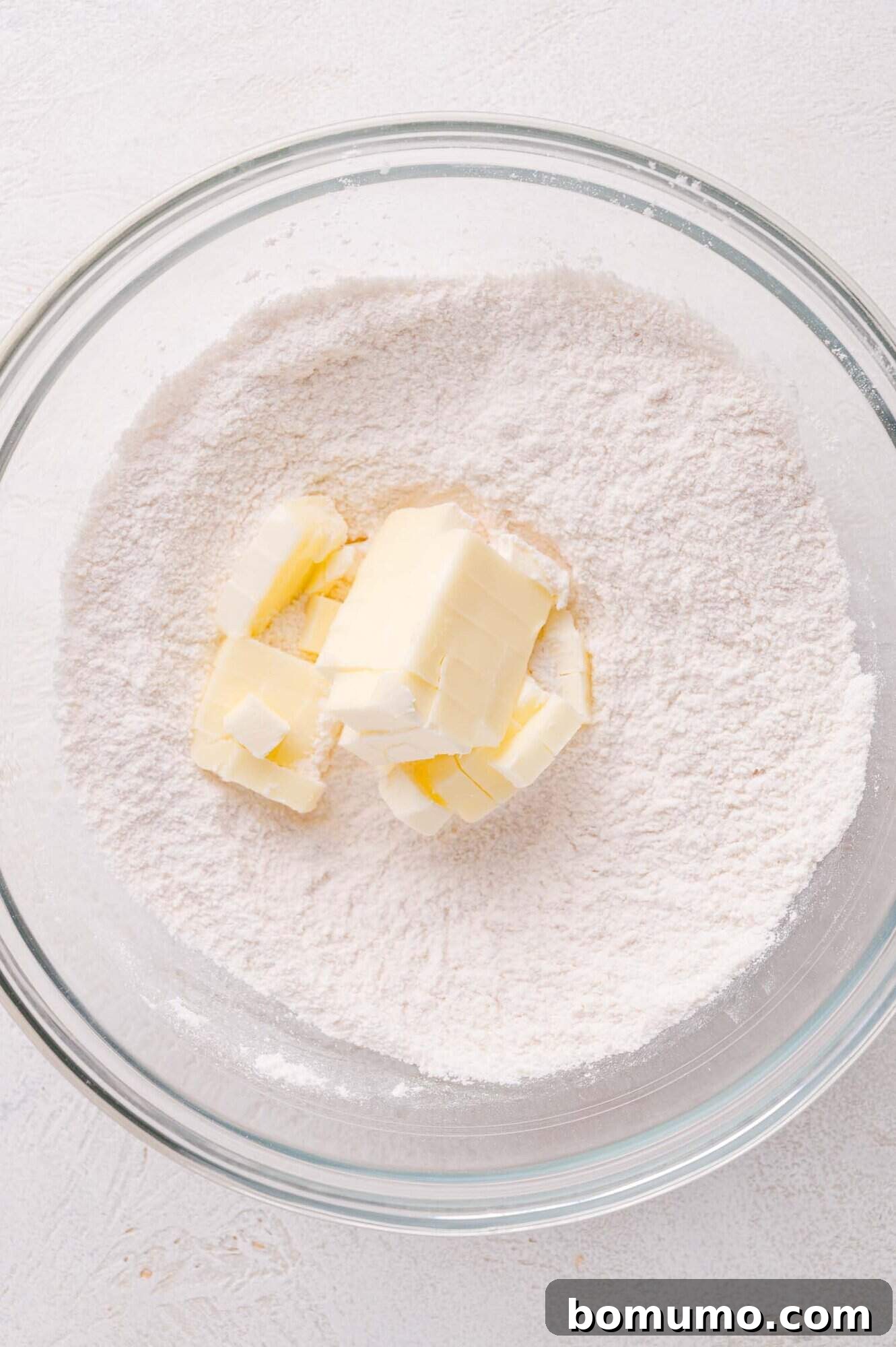 A top-down view of a clear glass bowl, containing a base of flour with precise cubes of cold butter strategically placed on top, poised for the next step in baking.