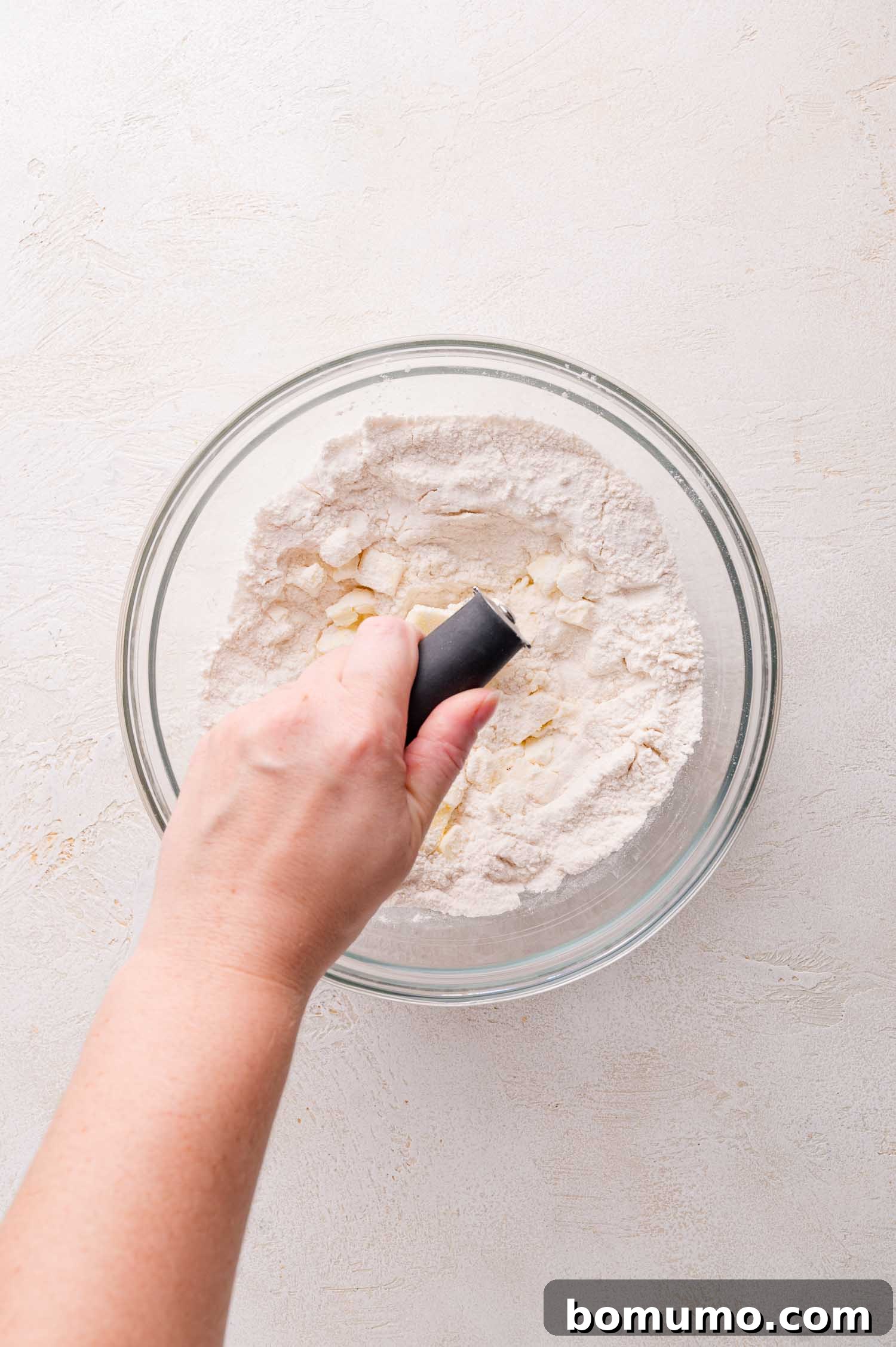A hand skillfully uses a pastry cutter to incorporate cold butter into the flour mixture within a glass mixing bowl, resting on a light-colored surface.