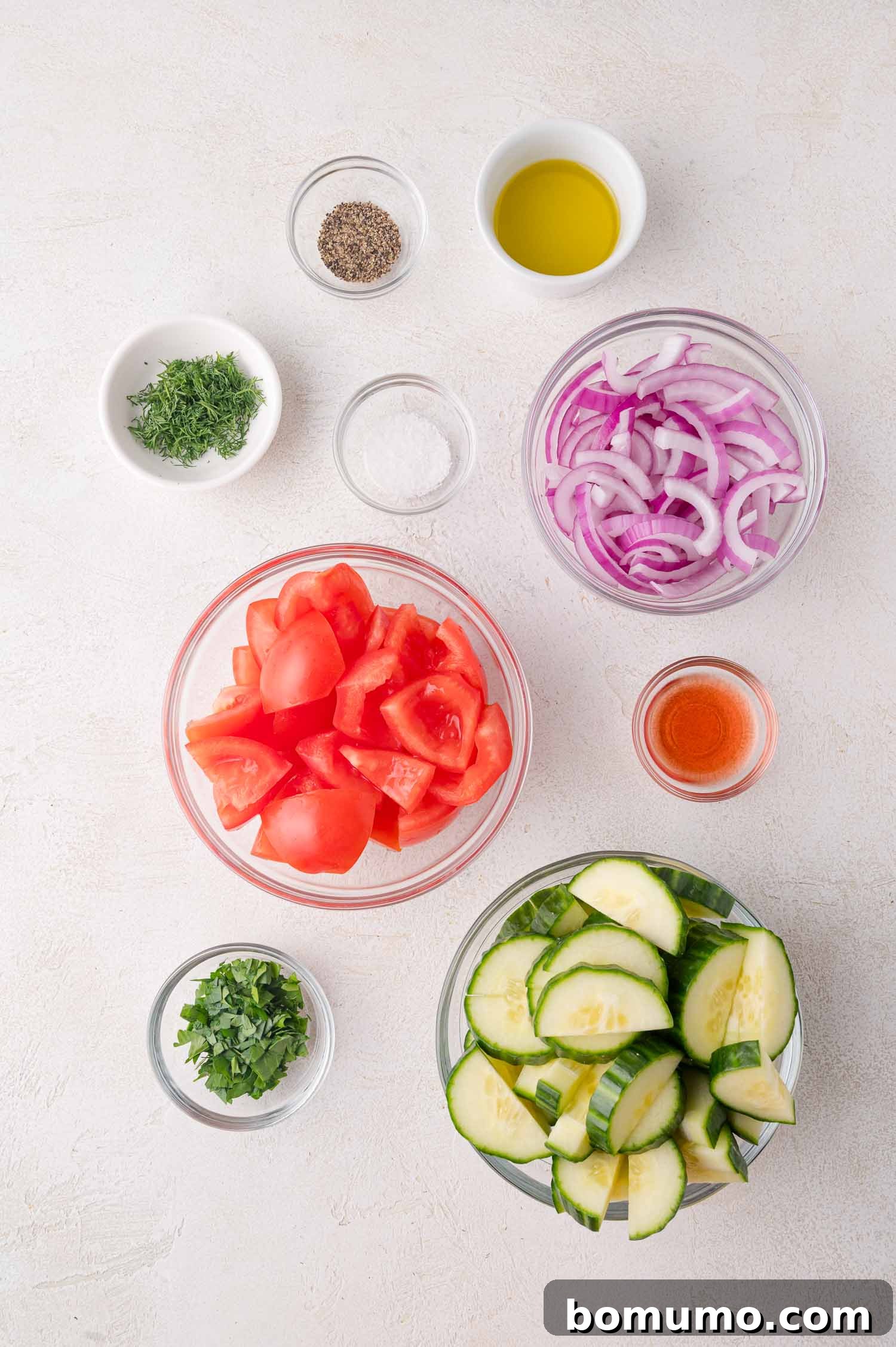 Bowls of chopped tomatoes, sliced cucumbers, sliced red onions, chopped herbs, and small bowls of olive oil, vinegar, salt, and pepper arranged on a white surface.
