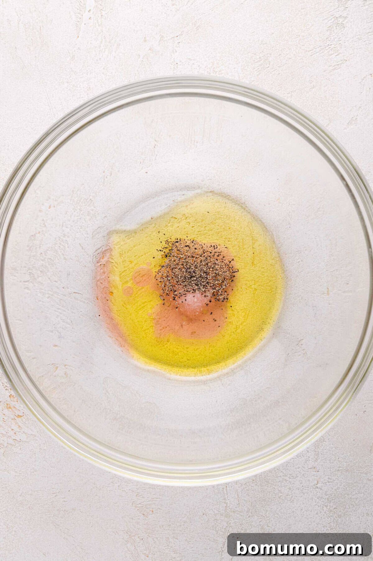 A glass mixing bowl containing olive oil, vinegar, and ground black pepper on a light-colored surface.