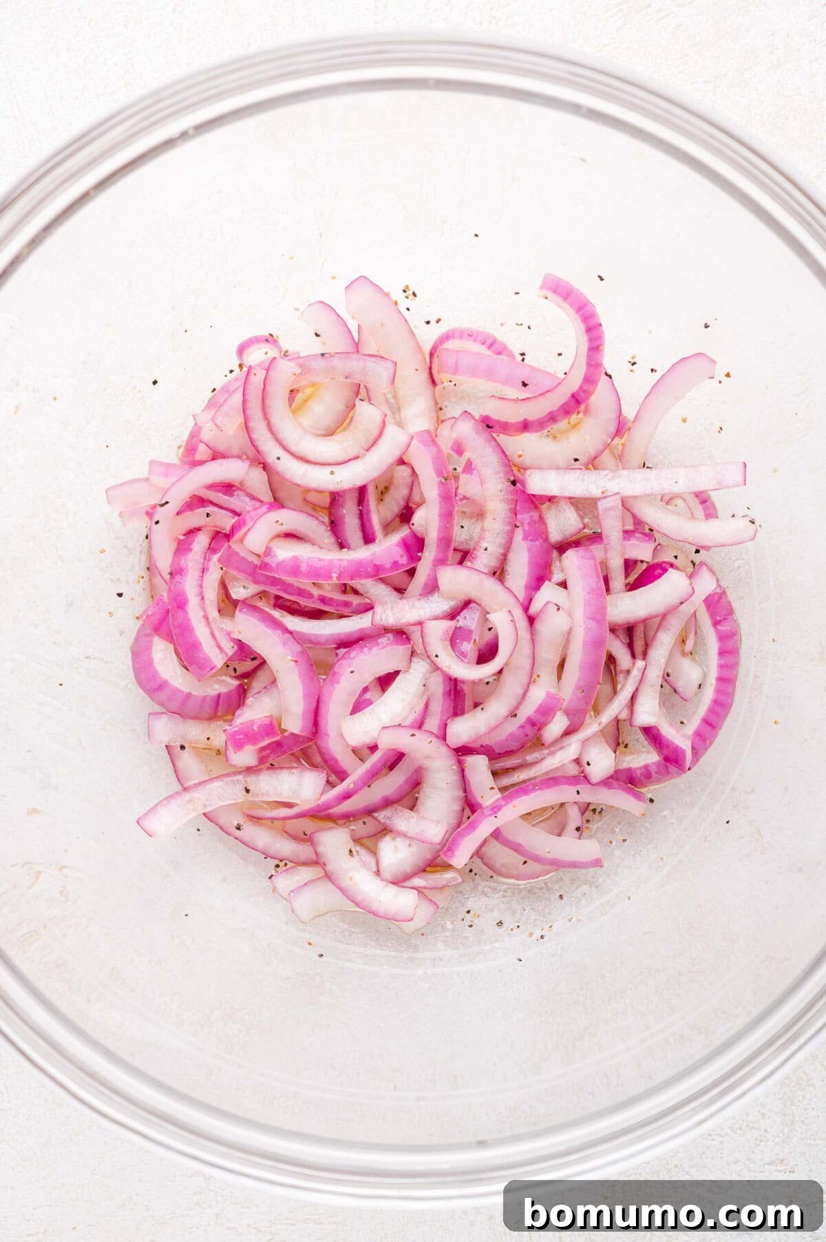 Sliced red onions with black pepper in a clear glass bowl on a light surface.