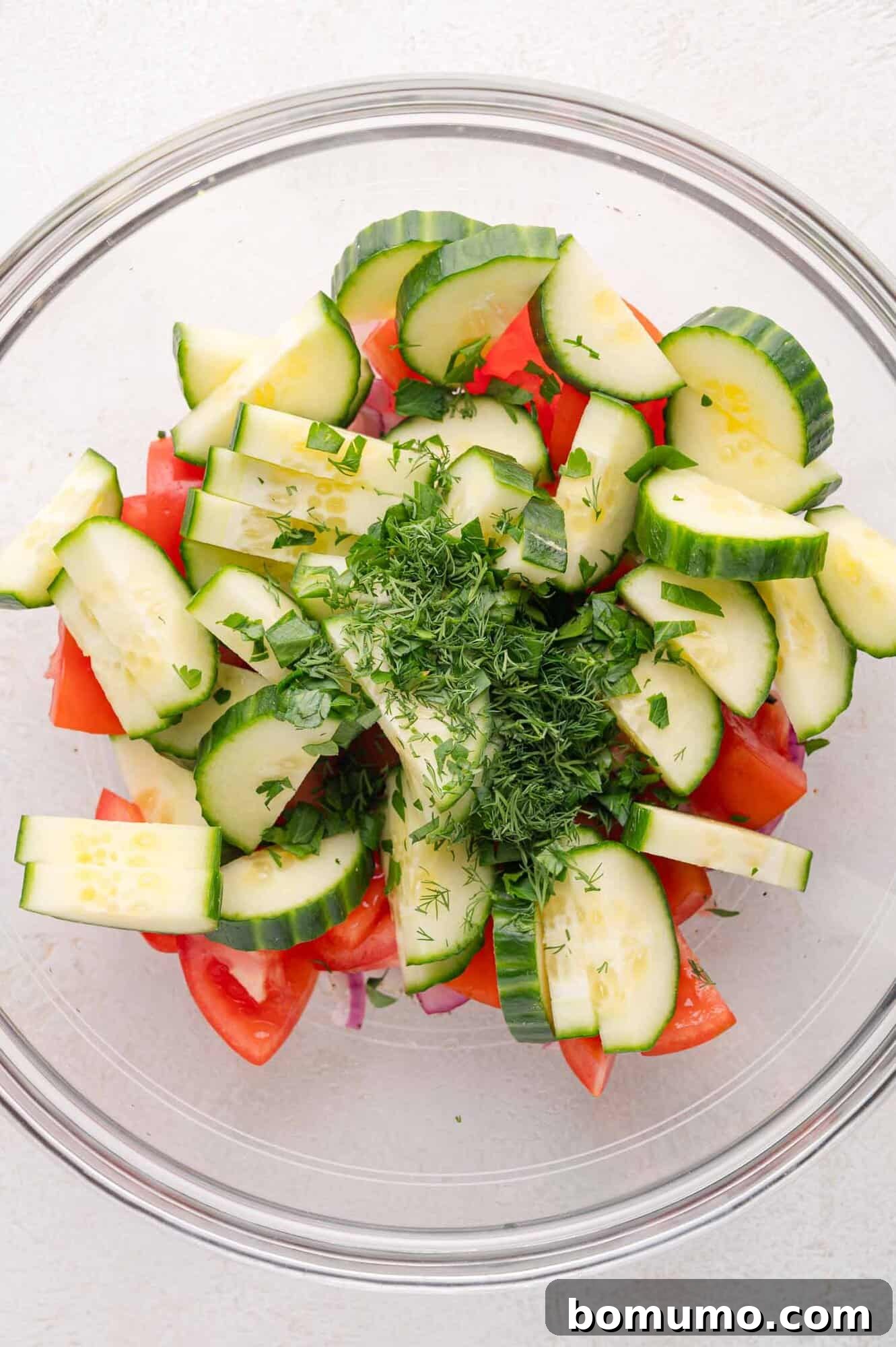 A glass bowl filled with sliced cucumbers, chopped tomatoes, red onions, and fresh chopped herbs on a light surface.