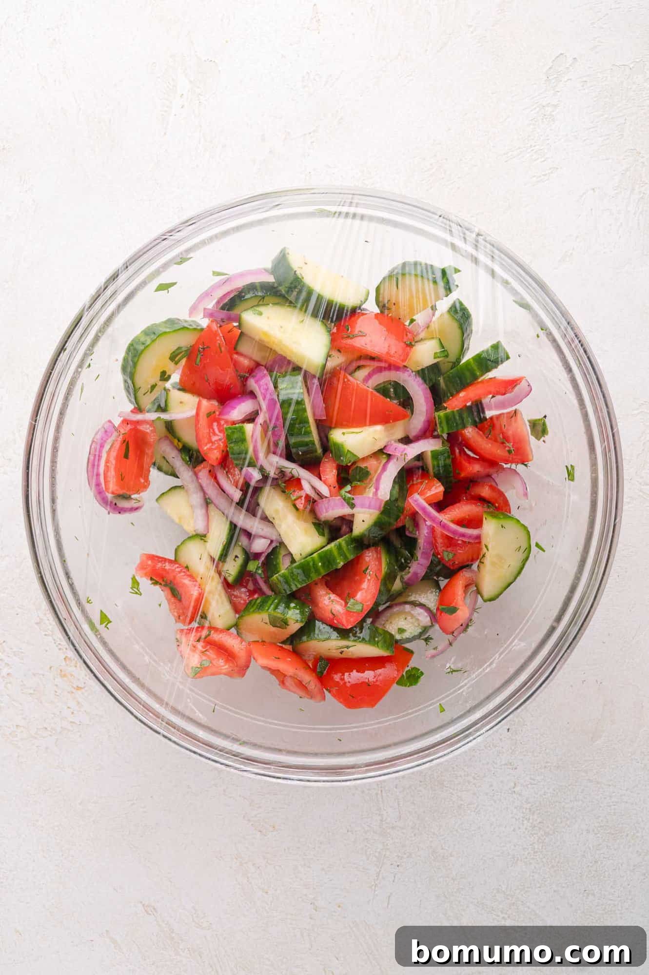 A glass bowl of cucumber, tomato, and red onion salad covered with plastic wrap, viewed from above on a light textured surface.