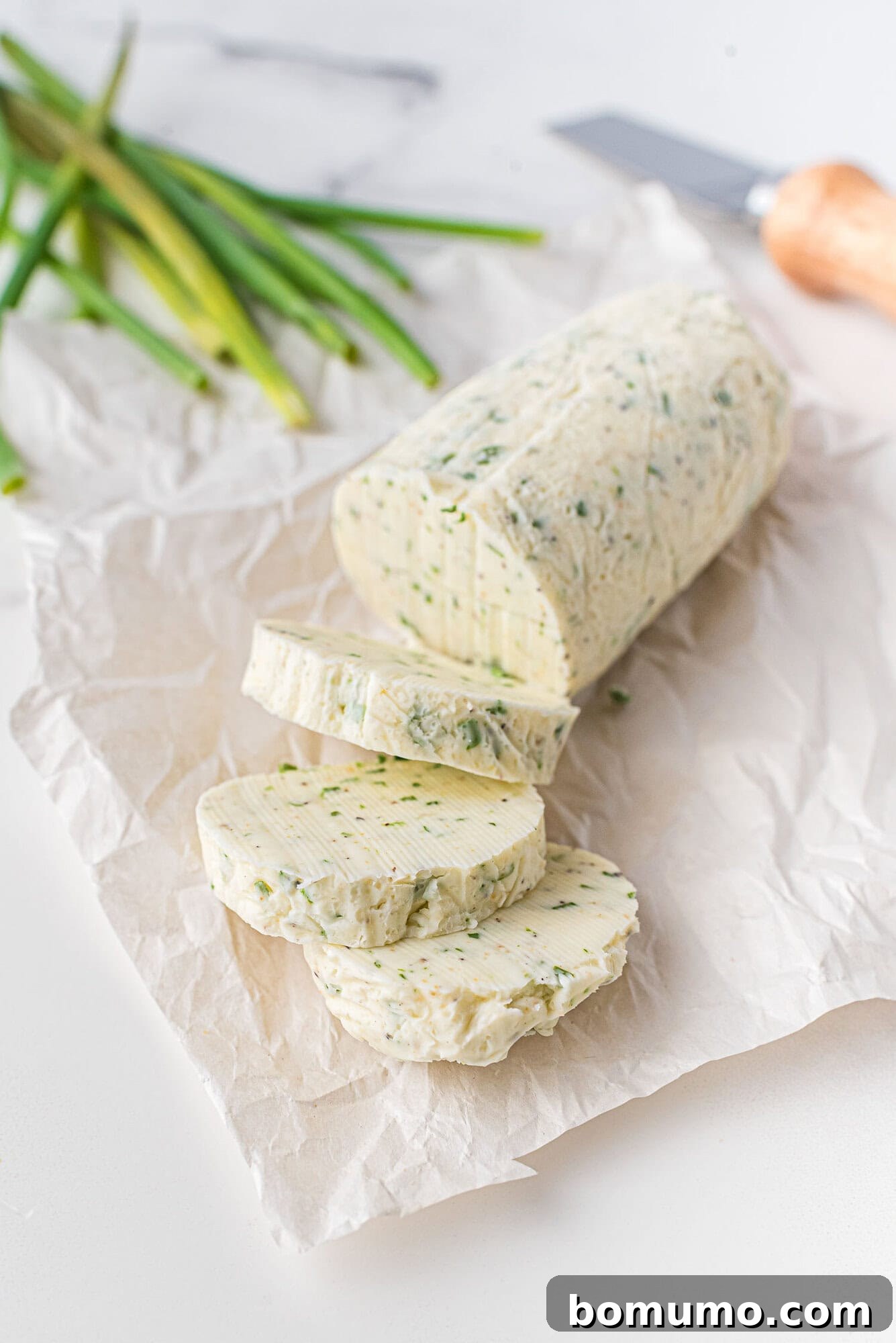 Verdant Chive Butter 2 A beautifully crafted log of chive butter, partially sliced on parchment paper, ready to be served. A knife and fresh chives lie in the background, hinting at the simple ingredients.