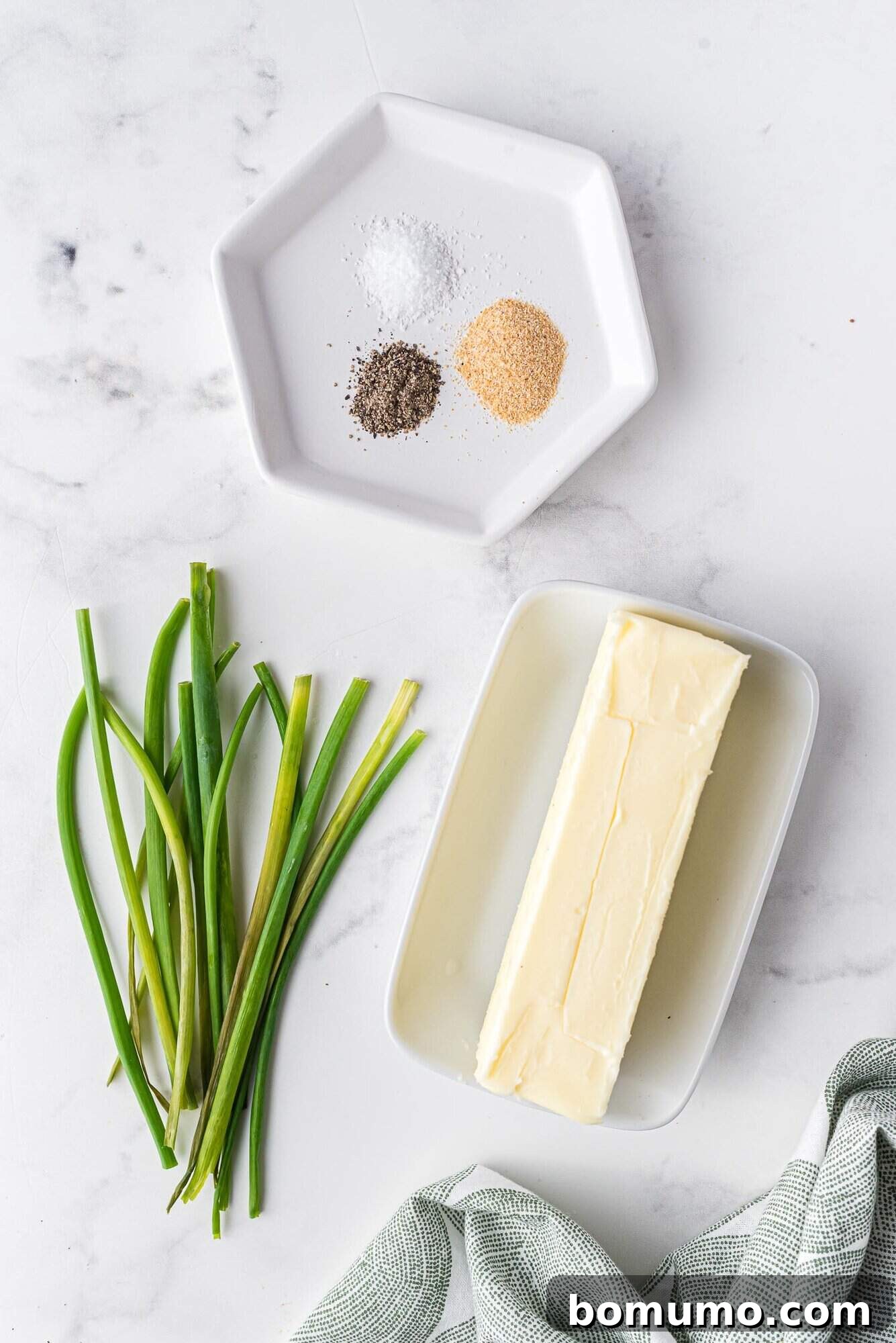 Verdant Chive Butter 3 A stick of softened butter, fresh chives, and a small plate with garlic powder, salt, and pepper on a pristine white marble surface, ingredients ready for mixing.