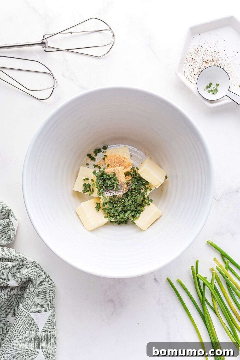 Verdant Chive Butter 4 A large white mixing bowl filled with chunks of room-temperature butter, finely chopped chives, garlic powder, salt, and pepper, set on a white countertop with a whisk nearby.
