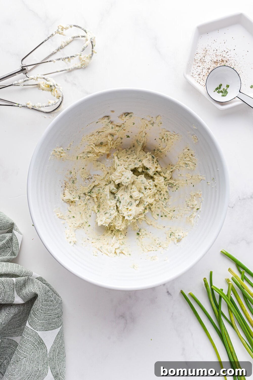 Verdant Chive Butter 5 A white bowl holds a perfectly whipped mixture of chive butter, garnished with a sprinkle of fresh chives. A hand mixer and spices are visible in the background, showcasing the mixing process.