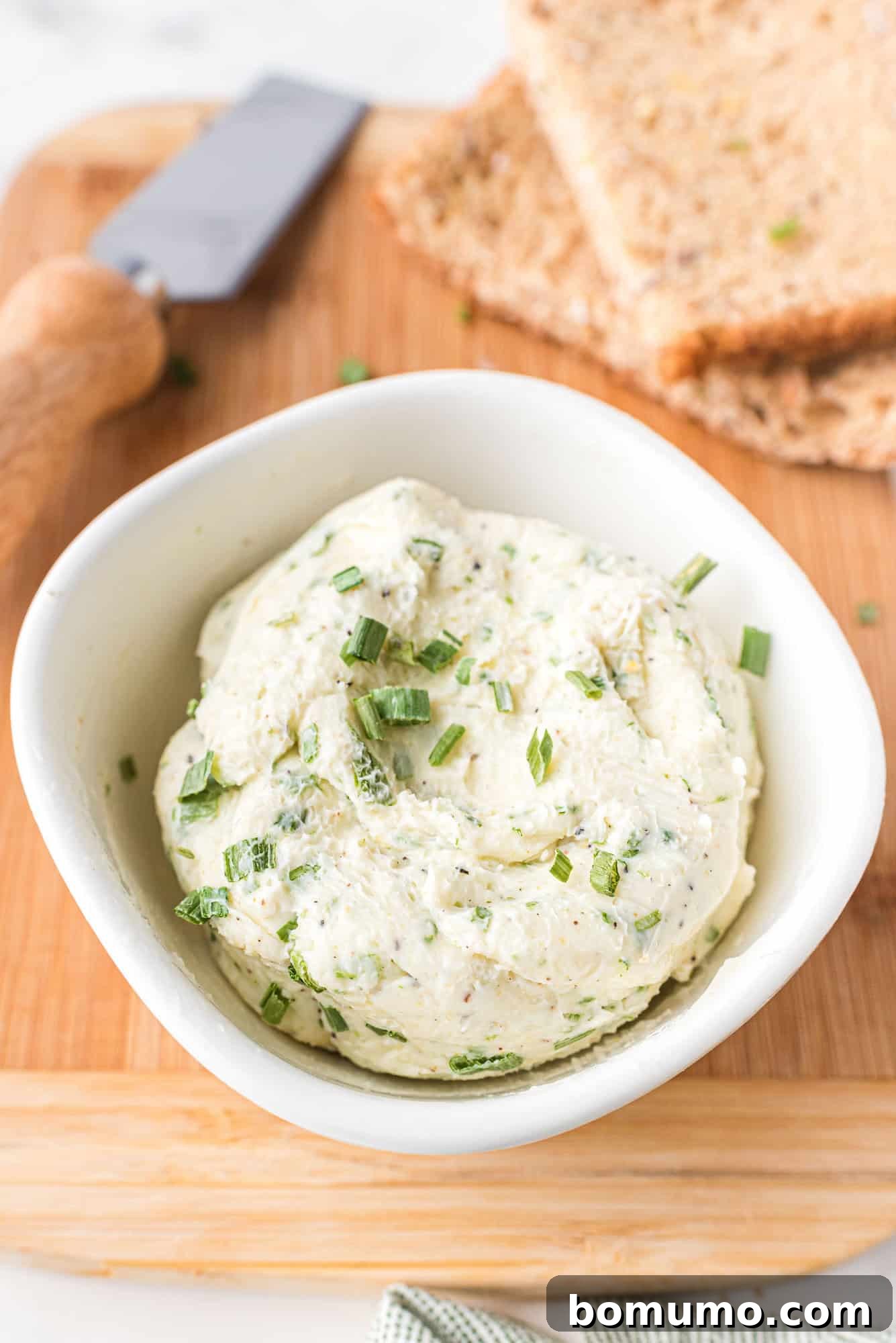 Verdant Chive Butter 6 A serving of freshly made chive butter in a small white bowl, adorned with extra chives, resting on a rustic wooden board alongside slices of wholesome whole grain bread and a butter knife.