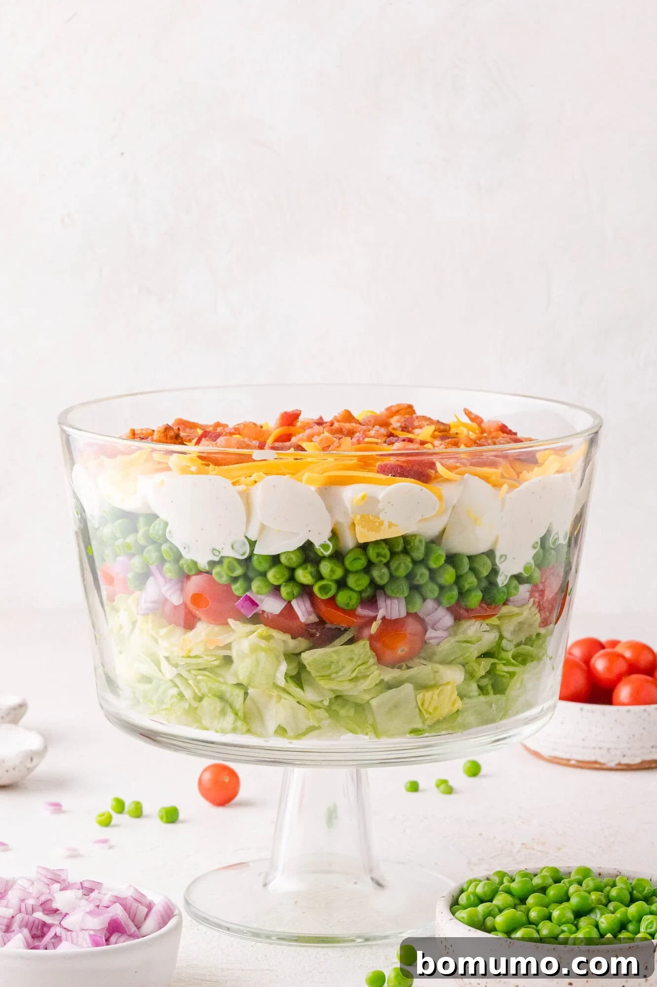 Stacked Garden Bliss 2 A beautiful glass trifle bowl showcasing a meticulously arranged 7 layer salad with vibrant layers of crisp lettuce, sweet onions, juicy cherry tomatoes, bright green peas, sliced hard-boiled eggs, fluffy shredded cheese, and crumbled crispy bacon.