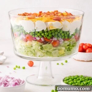 A glass trifle bowl filled with beautifully layered salad ingredients including crisp lettuce, sweet tomatoes, red onion, green peas, perfectly cooked eggs, rich cheese, and savory bacon.