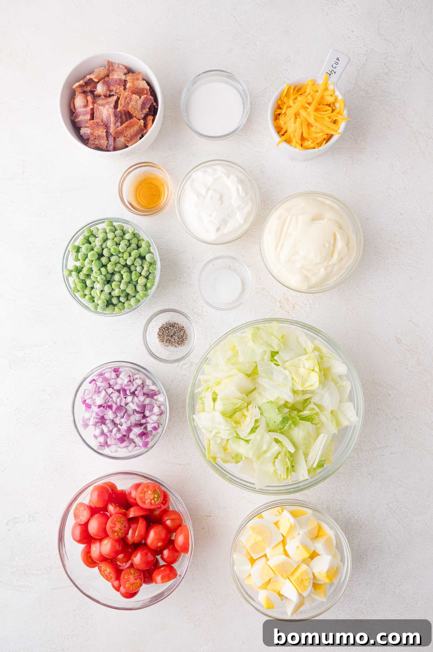 Stacked Garden Bliss 4 An overhead shot showcasing all the fresh and vibrant ingredients for the 7 layer salad, neatly arranged in separate bowls. Visible are crisp lettuce, halved cherry tomatoes, finely diced red onion, bright green frozen peas, sliced hard-boiled eggs, crumbled bacon, shredded cheddar cheese, and the essential dressing ingredients: mayonnaise, sour cream, vinegar, sugar, salt, and pepper.