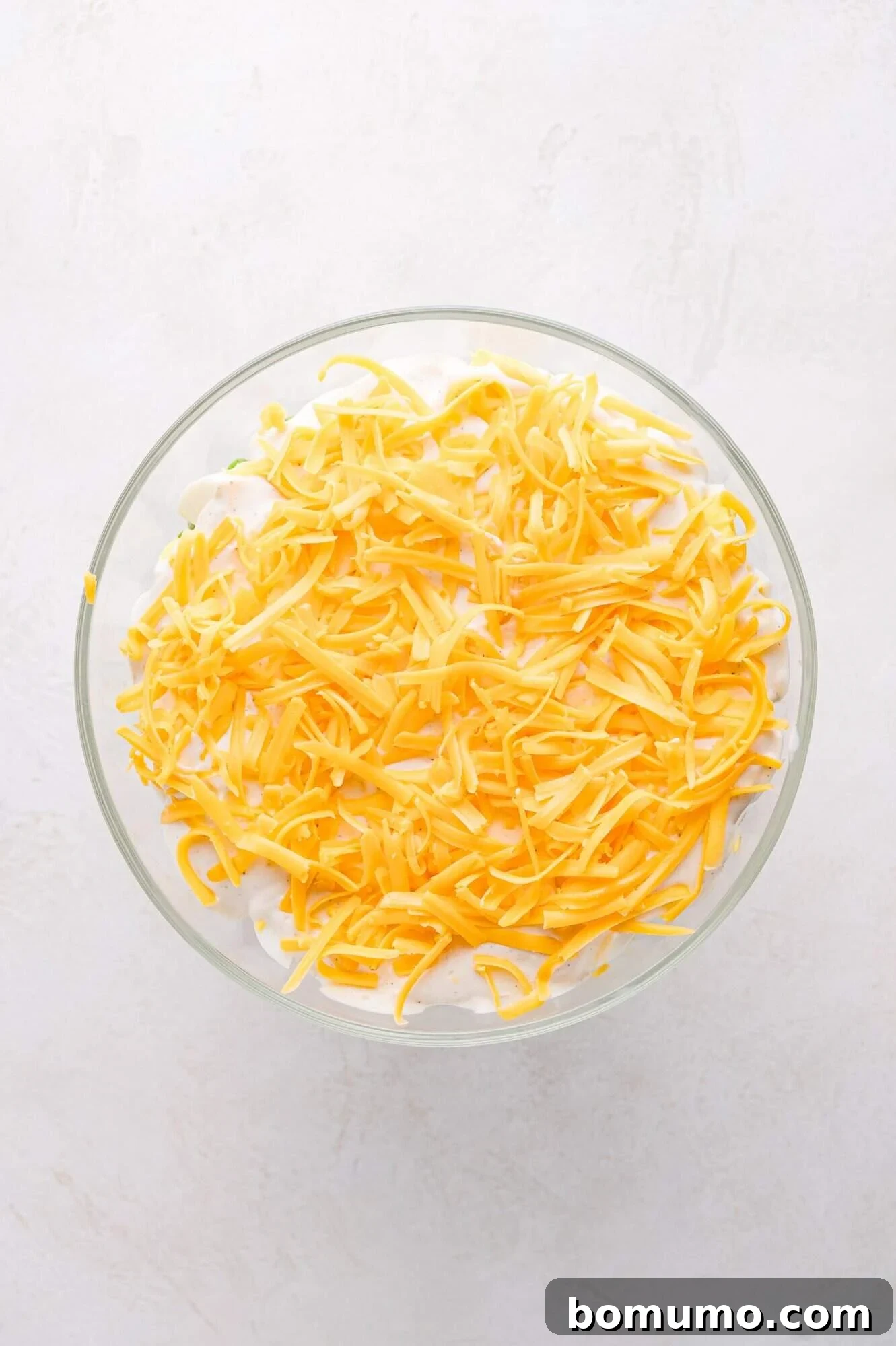 Stacked Garden Bliss 7 A top-down view of the 7 layer salad in a glass bowl, showcasing the top layer of bright shredded cheddar cheese, ready for chilling.