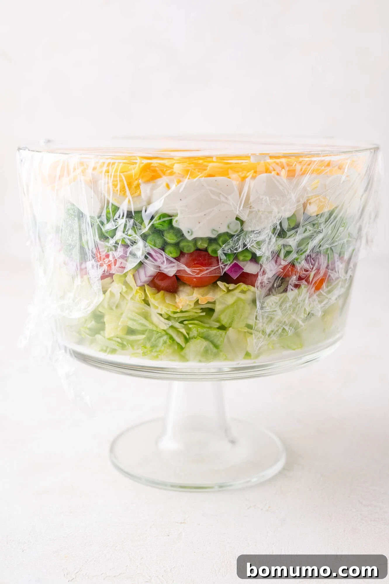 Stacked Garden Bliss 8 A glass trifle bowl elegantly filled with neatly layered salad ingredients, perfectly covered with plastic wrap, chilling in the refrigerator.