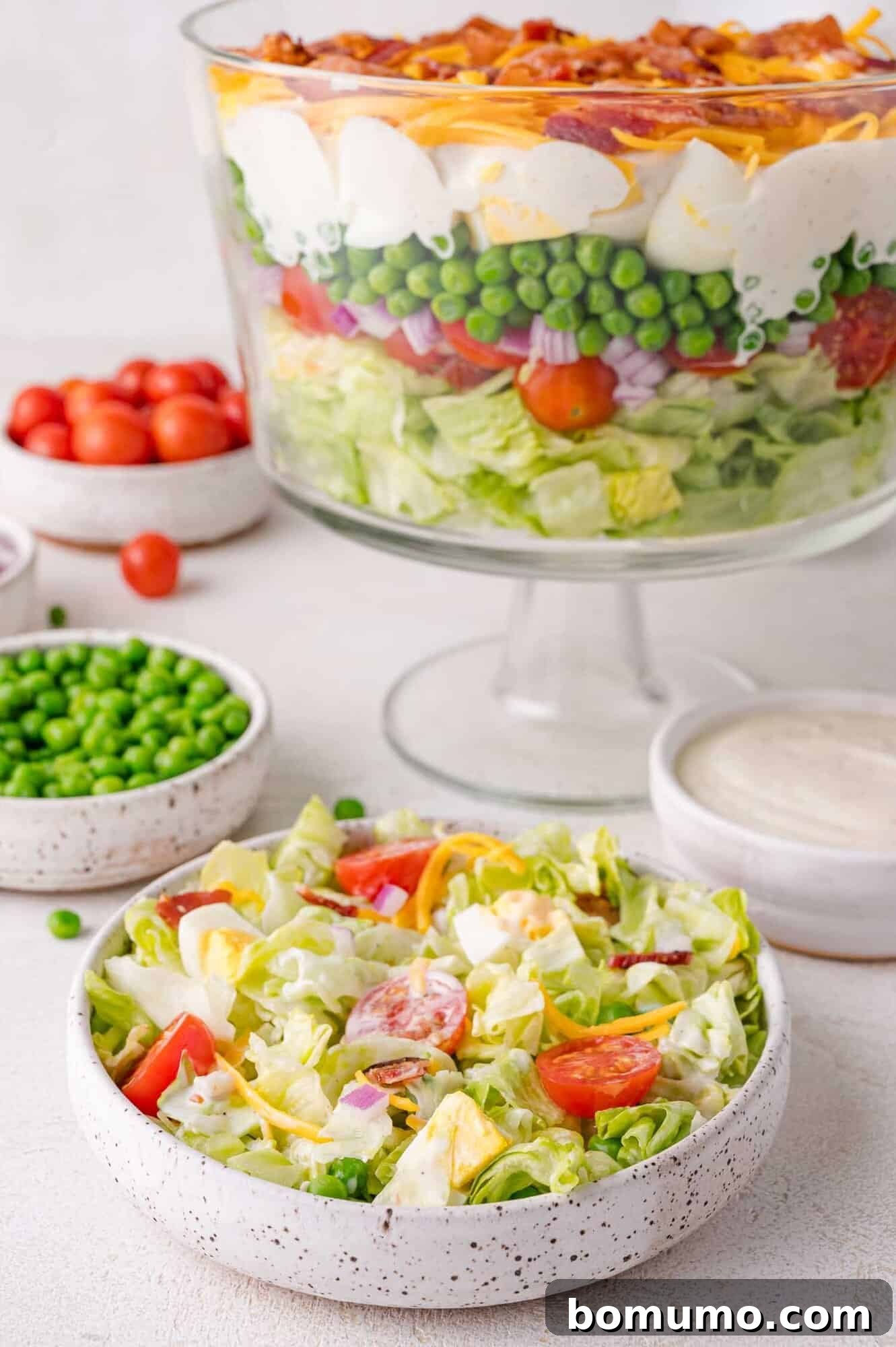 Stacked Garden Bliss 10 A grand bowl of 7 layer salad featuring distinct layers of lettuce, tomatoes, peas, eggs, cheese, and bacon, accompanied by a smaller serving bowl, dressing, and fresh ingredients.