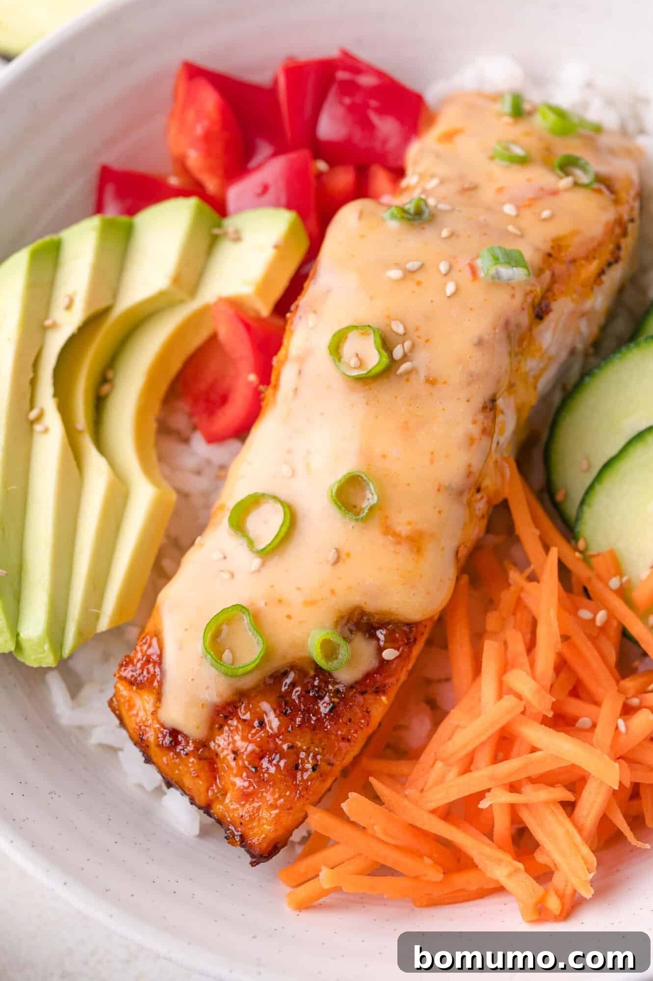 A vibrant bowl featuring fluffy white rice, neatly sliced avocado, ripe tomatoes, crisp cucumber, shredded carrots, and a perfectly baked piece of Bang Bang salmon, elegantly garnished with sesame seeds and fresh green onions.