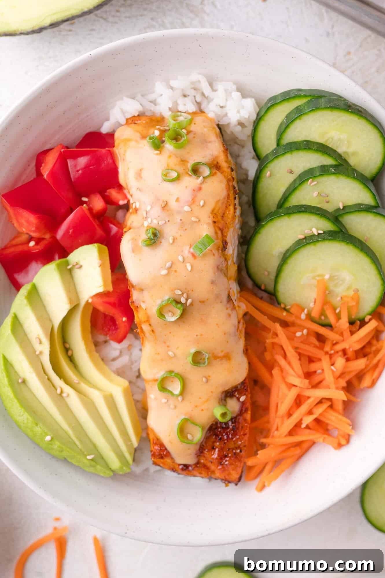 A vibrant and appealing bowl featuring fluffy white rice, topped with a beautifully glazed Bang Bang salmon fillet, alongside fresh slices of cucumber, delicate shredded carrots, creamy avocado, diced red bell pepper, and garnished with aromatic sesame seeds and fresh green onion.