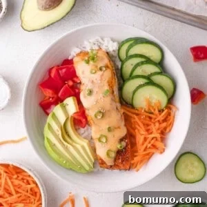 A bowl of white rice topped with bang bang salmon, sliced cucumber, avocado, diced tomatoes, shredded carrots, and a creamy sauce, garnished with green onions and sesame seeds.