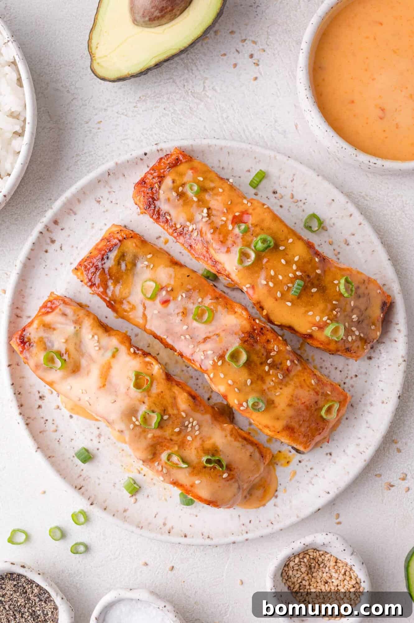 A speckled plate showcases three perfectly glazed Bang Bang salmon pieces, garnished with vibrant sliced green onions and toasted sesame seeds, artfully arranged alongside small bowls of sauce, fluffy rice, various seasonings, and half an avocado.