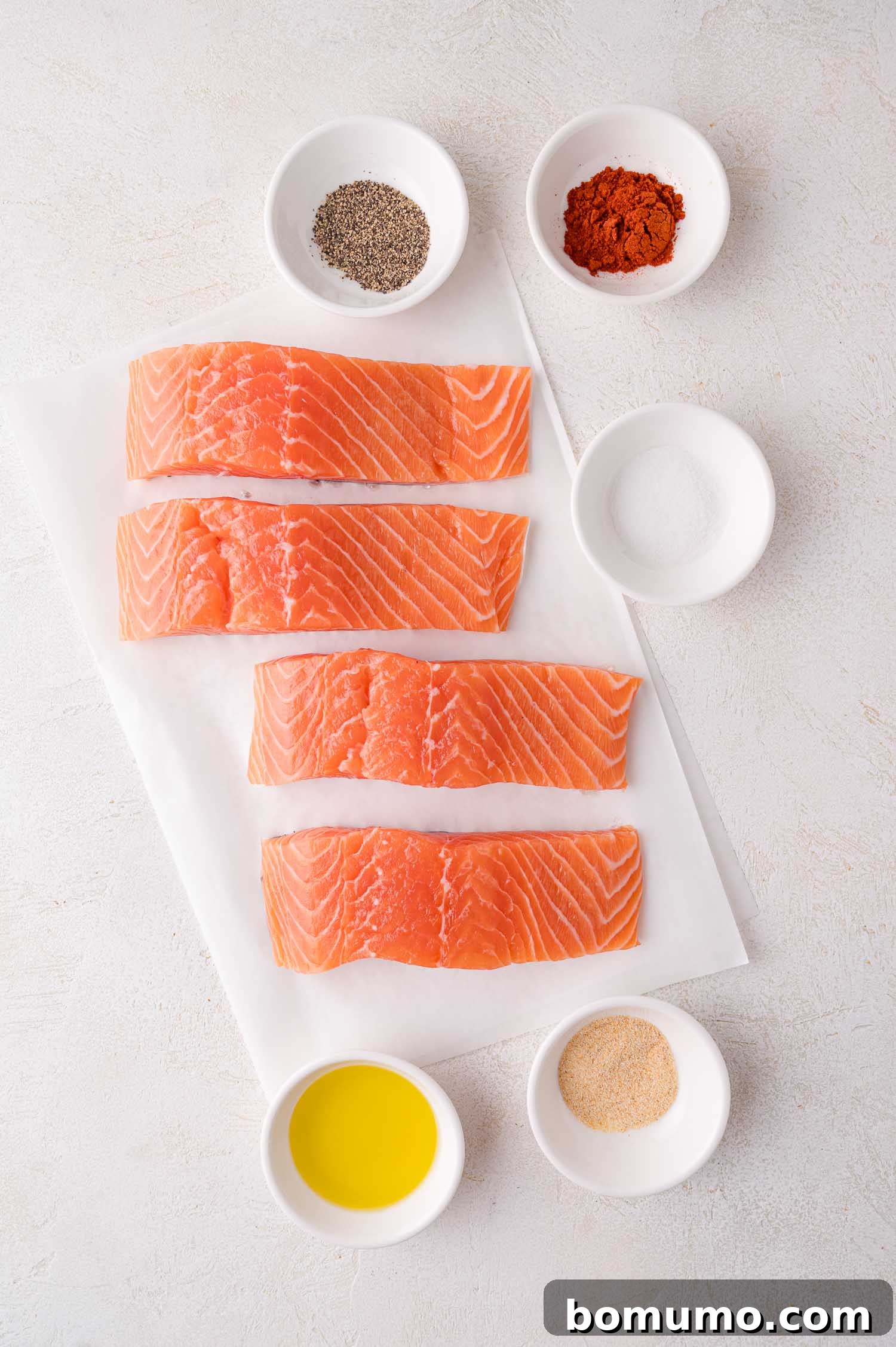 Four pristine raw salmon fillets rest on parchment paper, surrounded by small white bowls containing individual spices: black pepper, vibrant paprika, coarse salt, golden olive oil, and aromatic garlic powder, all meticulously arranged for seasoning.