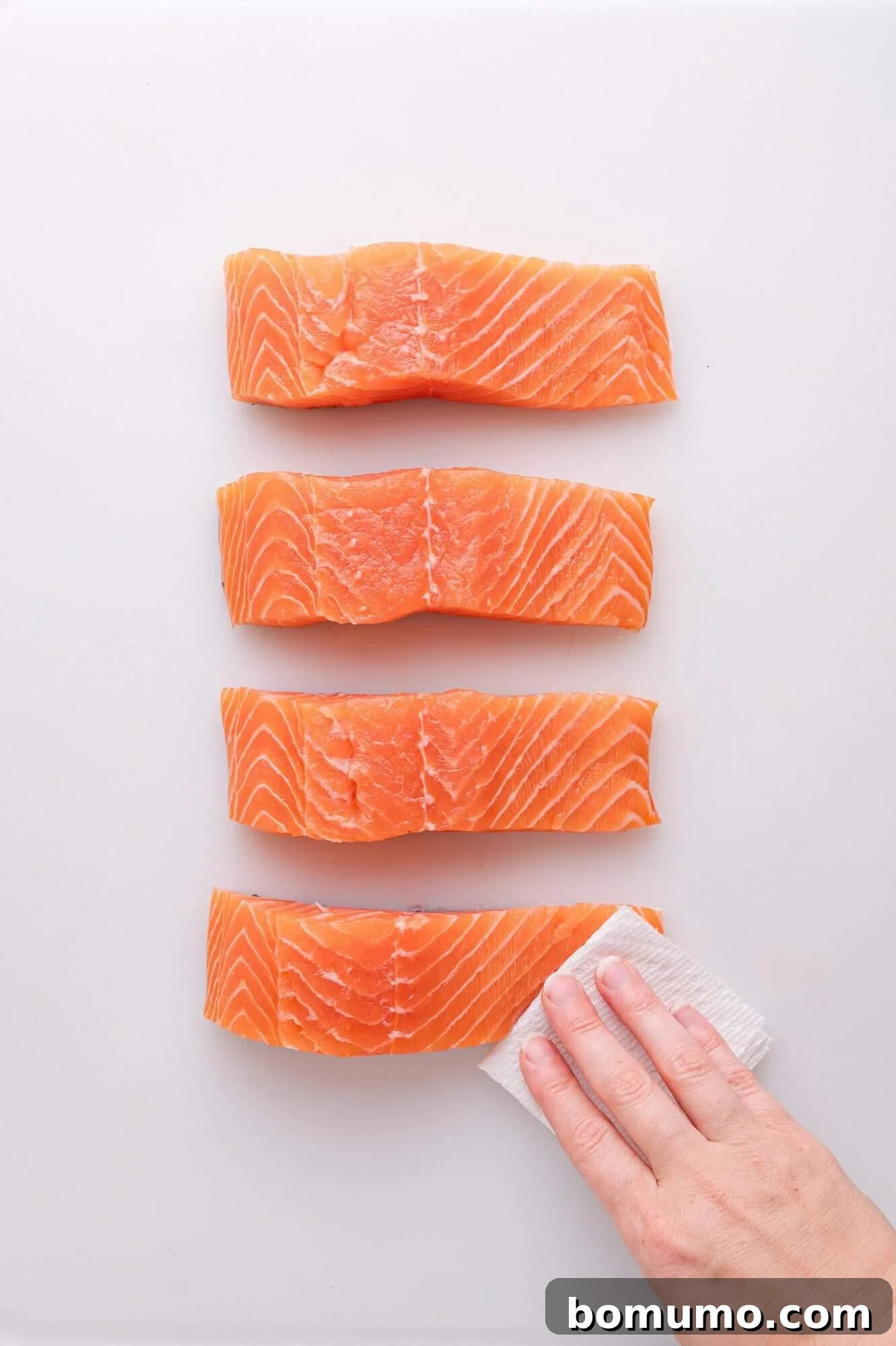 A hand gently pats one of four raw salmon fillets dry with a paper towel, as the fillets are neatly arranged in a row on a clean white surface, showcasing the initial step in preparation.