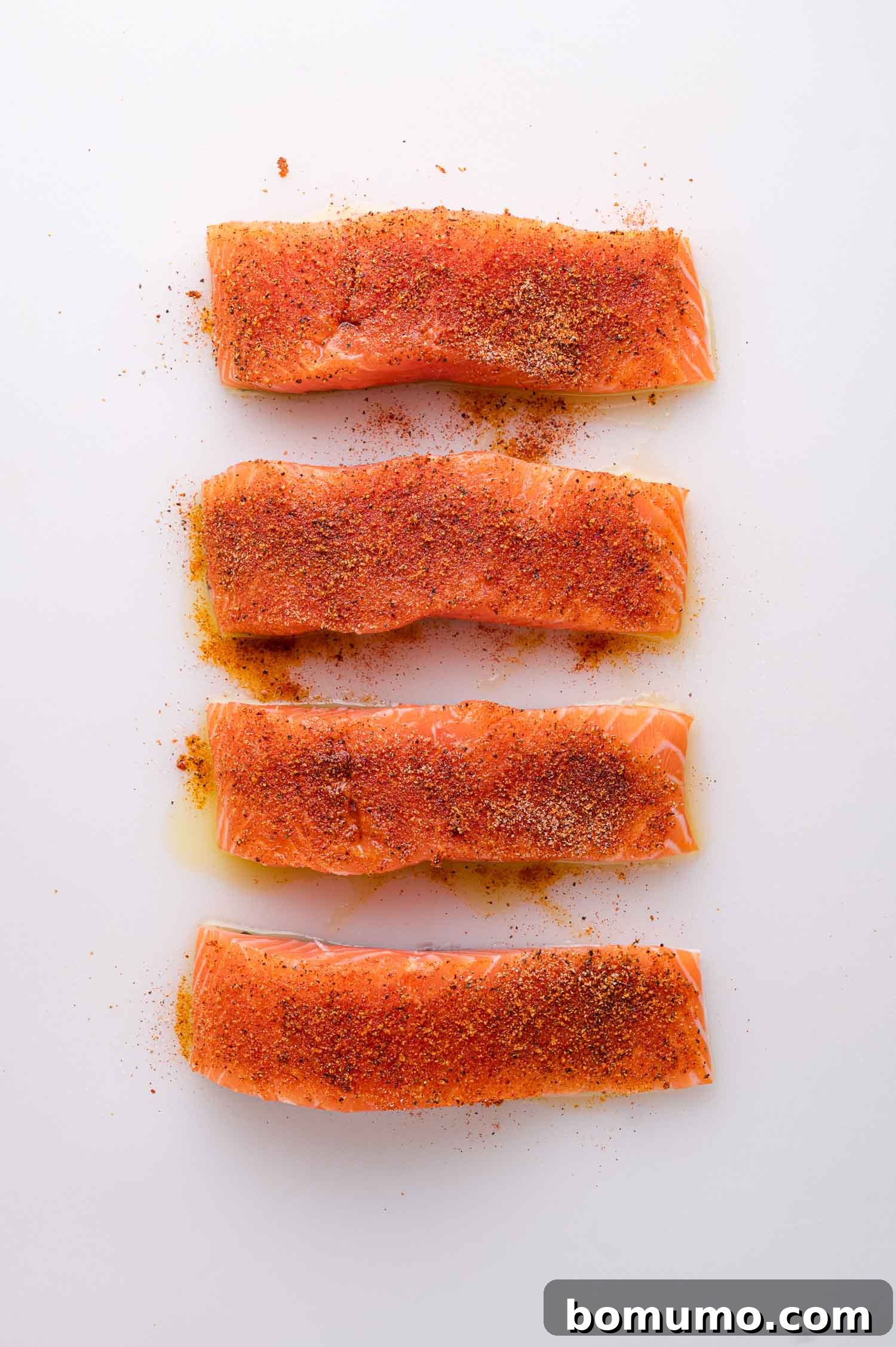 Four raw salmon fillets are meticulously arranged vertically on a pristine white surface, each generously topped with a rich, reddish-brown blend of aromatic spices, prepared for the next stage of cooking.