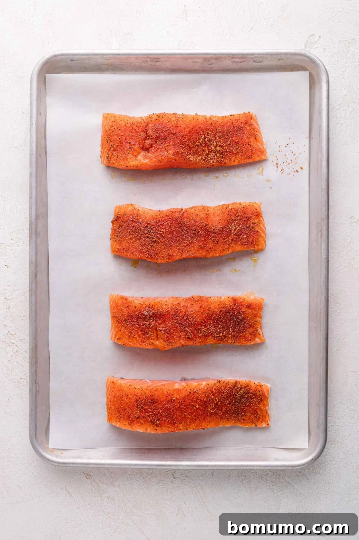 Four perfectly seasoned salmon fillets are neatly arranged in a single row on a parchment-lined baking sheet, awaiting their delicious Bang Bang sauce topping.