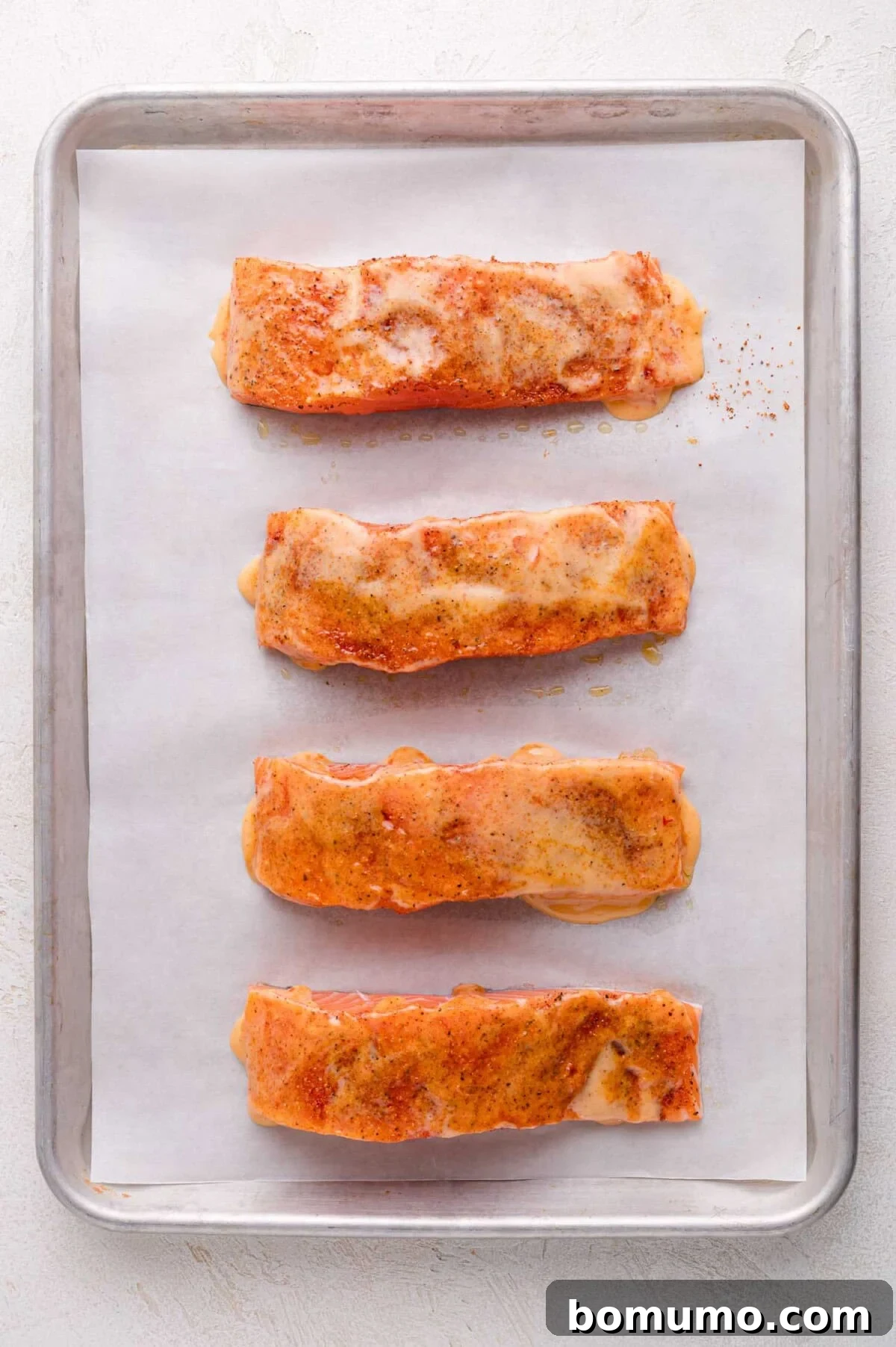 Four seasoned salmon fillets, now generously coated with a creamy sauce, are arranged in a single row on a parchment-lined baking sheet, perfectly prepped and ready for the oven.