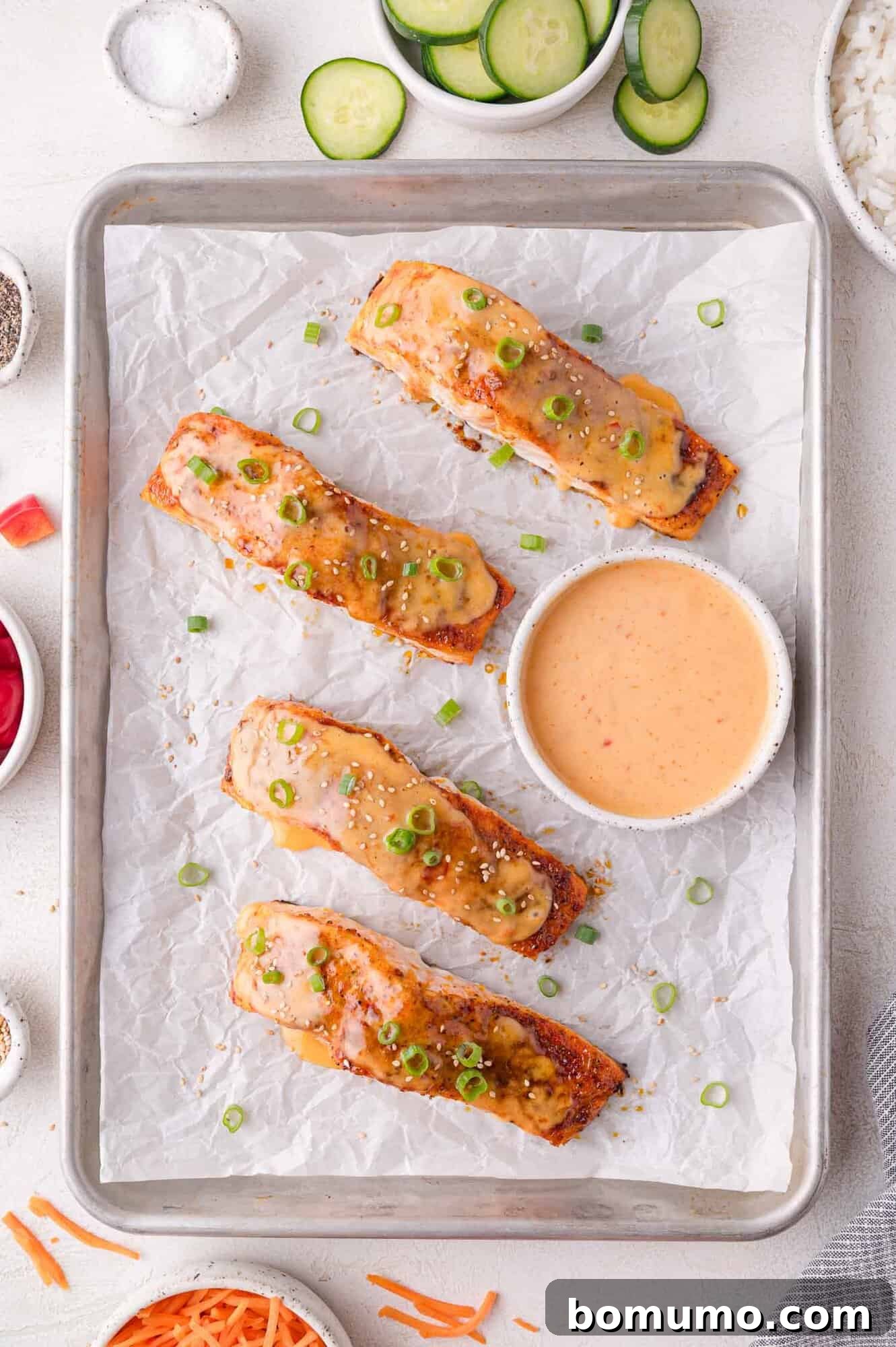 Four exquisitely baked salmon fillets, topped with a creamy sauce and vibrant chopped green onions, are presented on a parchment-lined baking sheet. A small bowl of extra dipping sauce sits in the center, flanked by bowls of refreshing sliced cucumbers and fluffy rice.