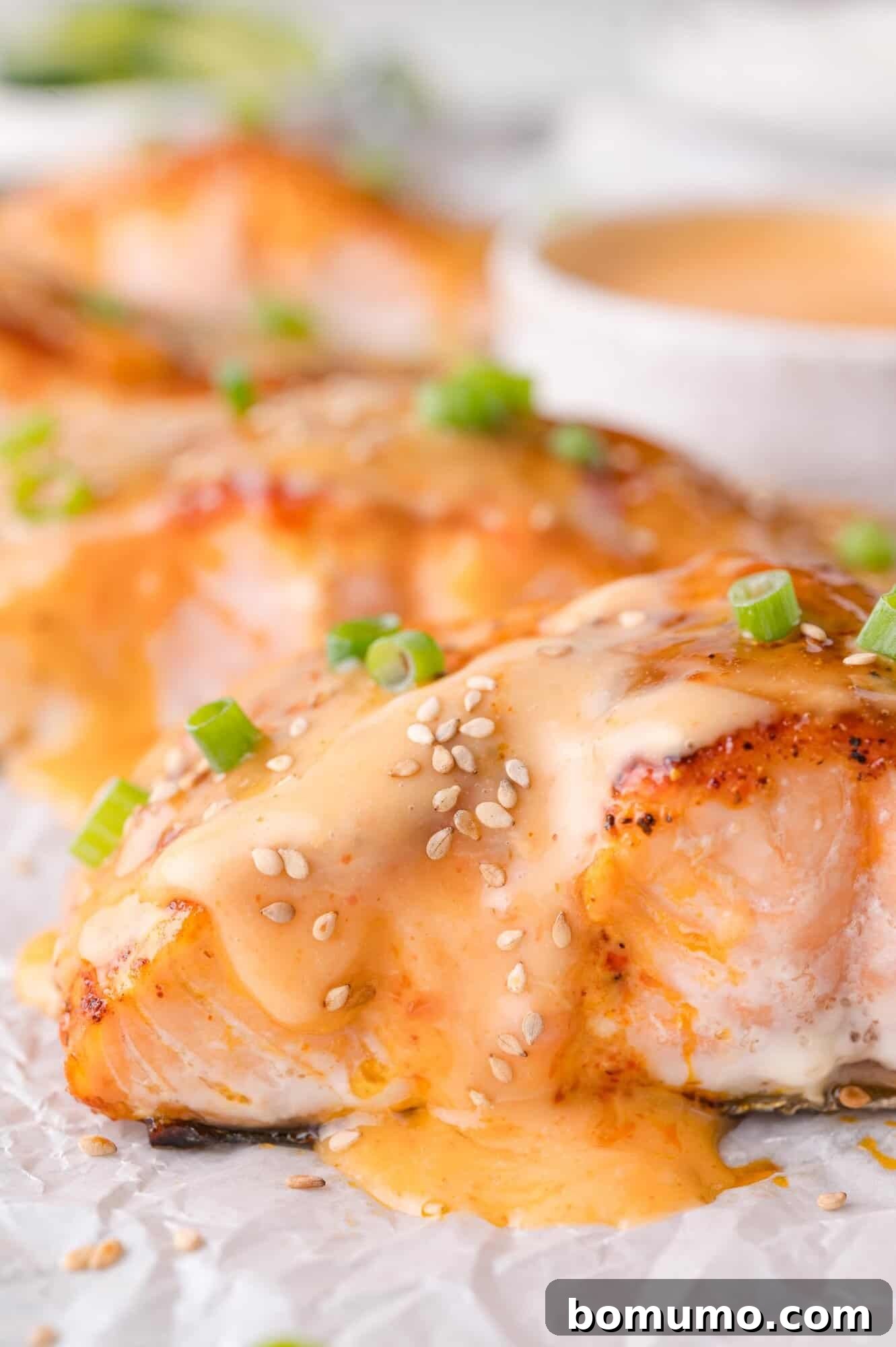 A close-up shot of perfectly cooked salmon fillets, glistening with creamy Bang Bang sauce and adorned with toasted sesame seeds and freshly chopped green onions, presented beautifully on parchment paper.