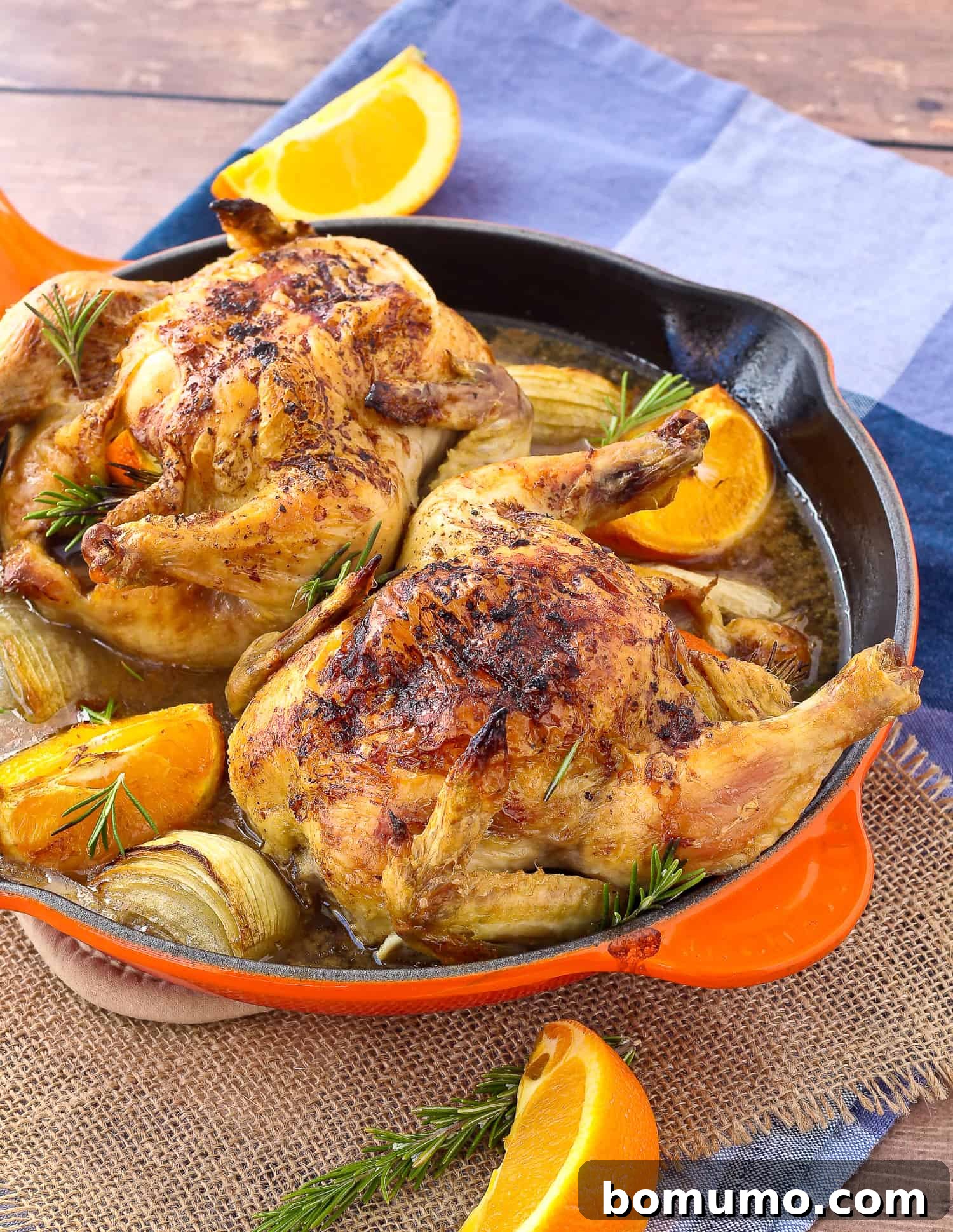 Two golden-brown Cornish game hens in a skillet, garnished with fresh orange slices and rosemary sprigs.