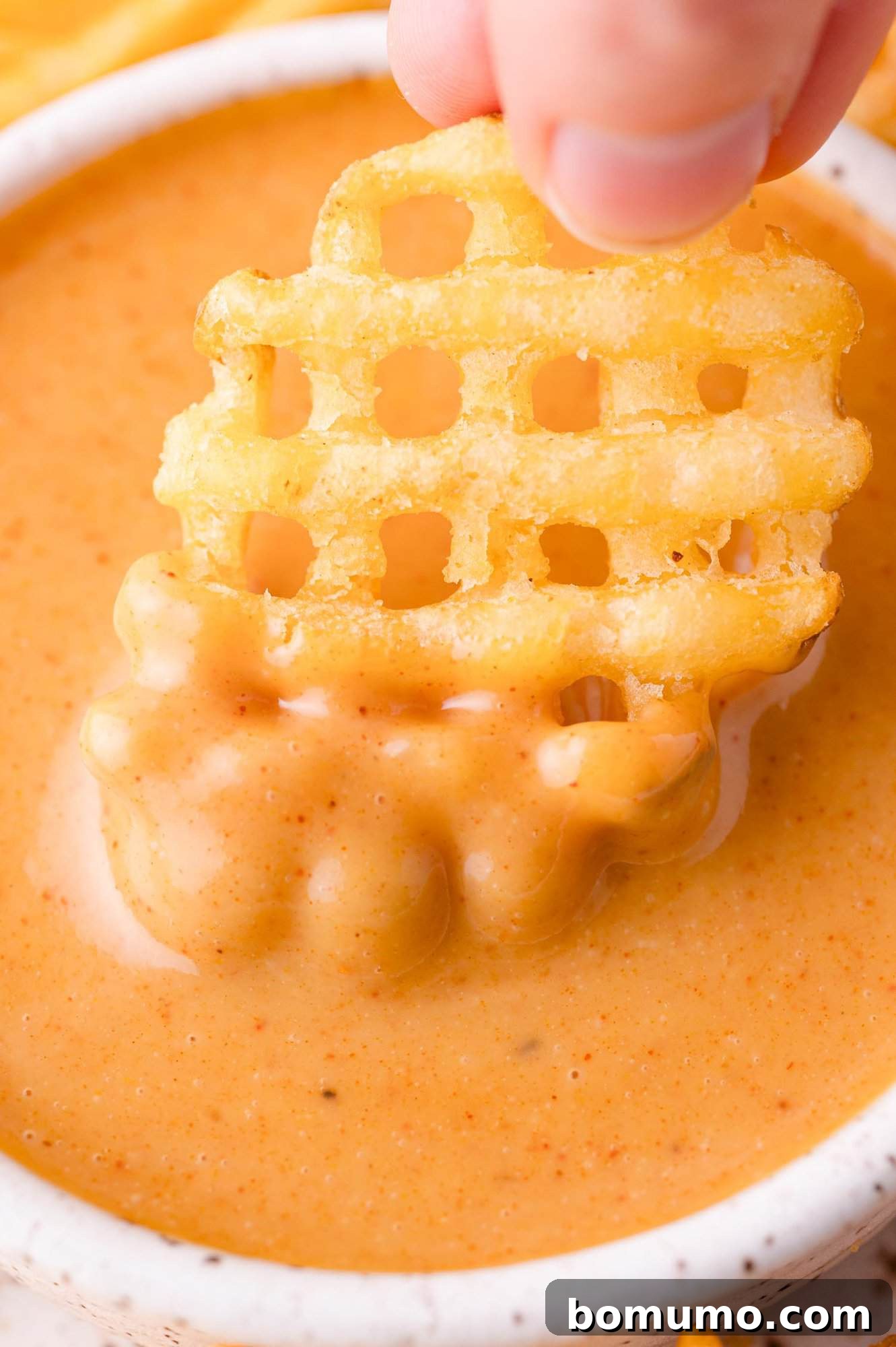 A hand dips a waffle-cut french fry into a bowl of creamy orange Chick-fil-A dipping sauce.