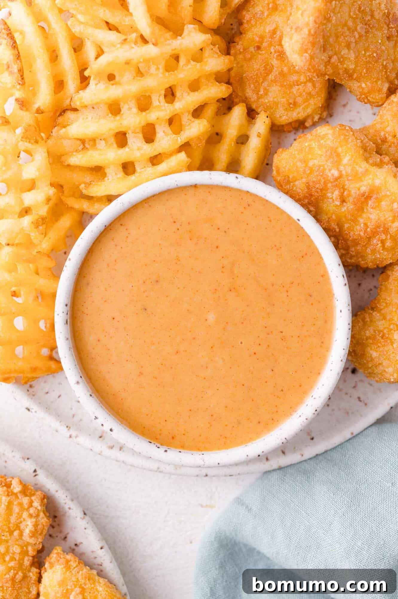 A bowl of creamy dipping sauce is surrounded by waffle fries and breaded chicken nuggets on a plate.