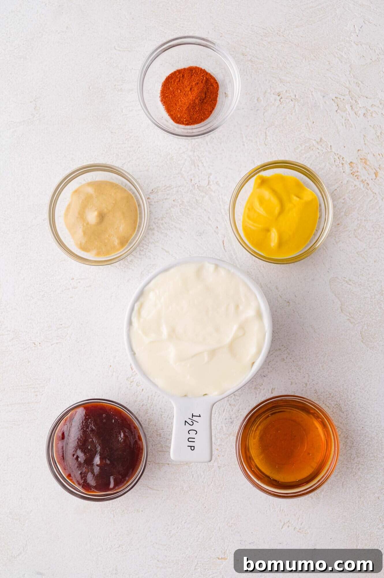 Six small containers with various condiments and spices, including mayonnaise, mustard, honey, barbecue sauce, Dijon mustard, and paprika, arranged on a light surface.