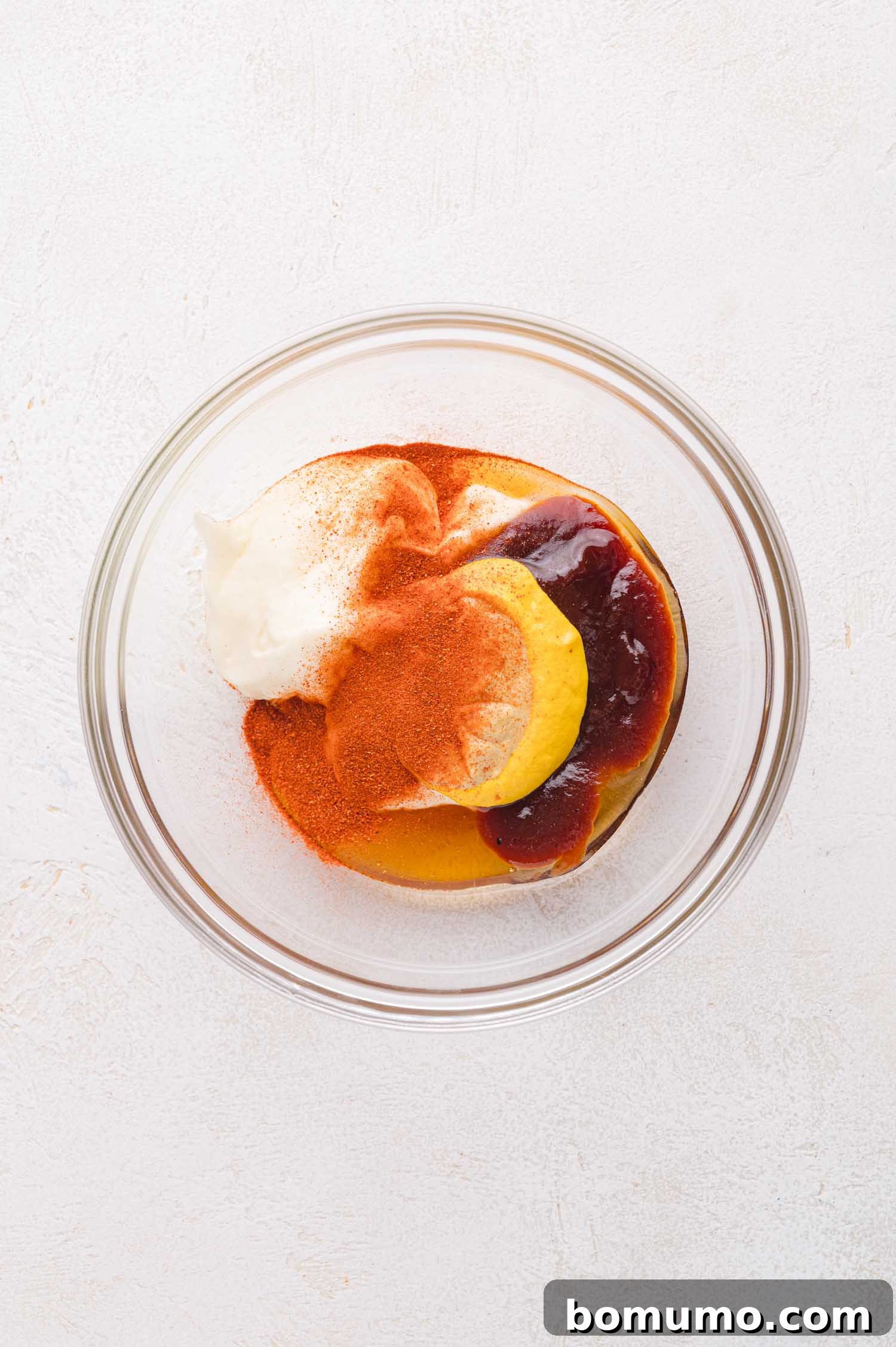 A glass bowl containing mayonnaise, mustard, barbecue sauce, and paprika on a white surface.