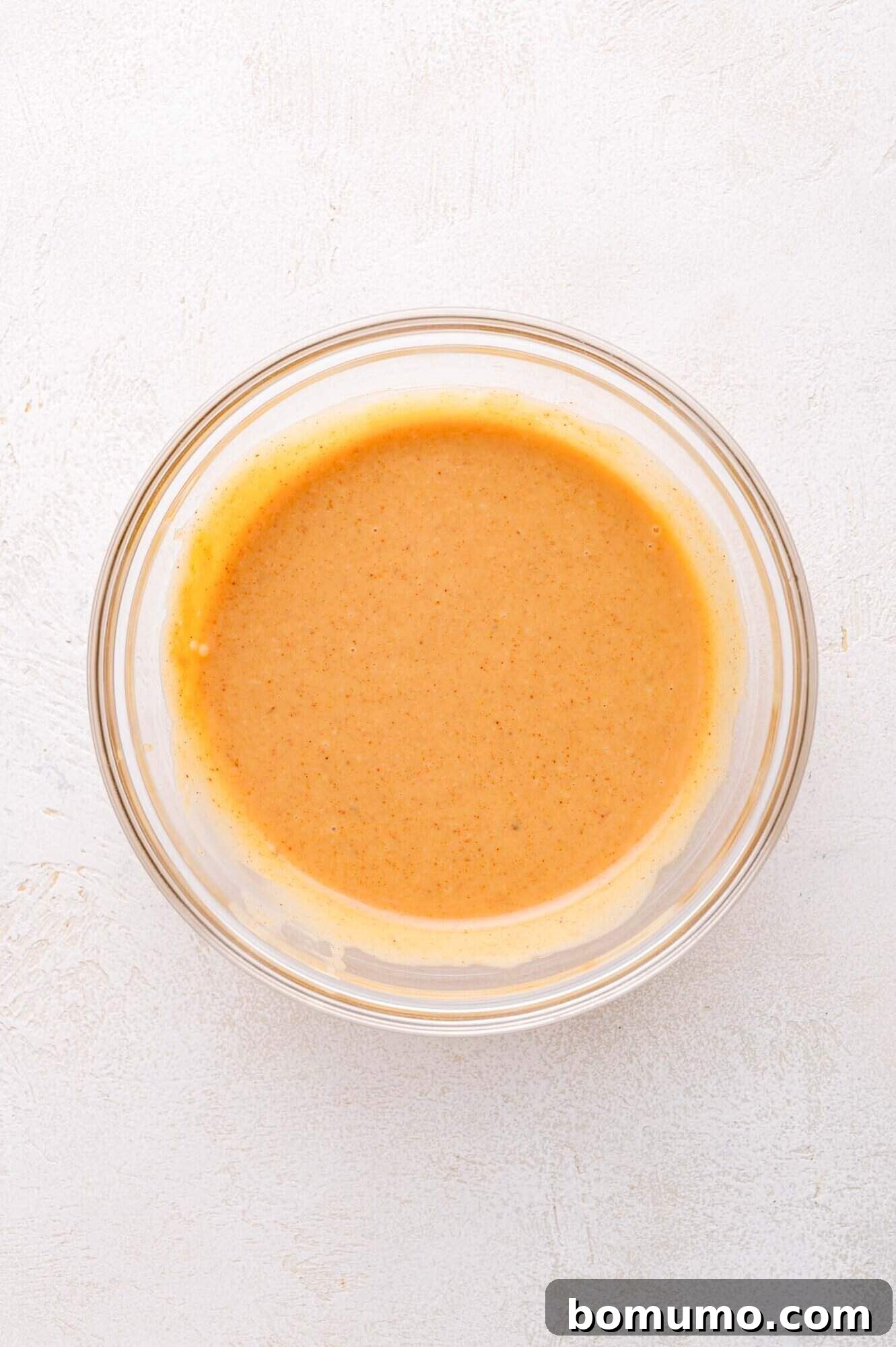 A clear glass bowl containing a smooth, light brown sauce or dressing sits on a white textured surface.