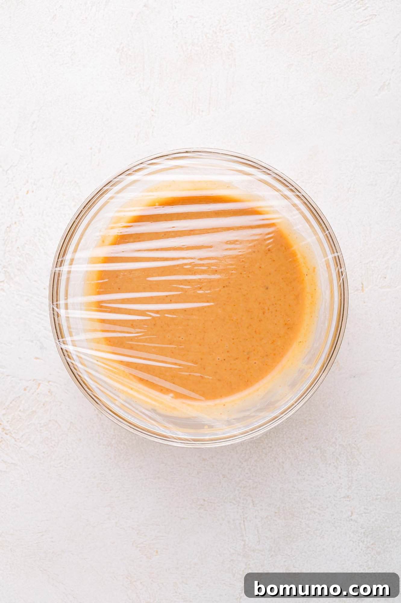A glass bowl filled with orange sauce is covered with plastic wrap and placed on a light-colored surface.