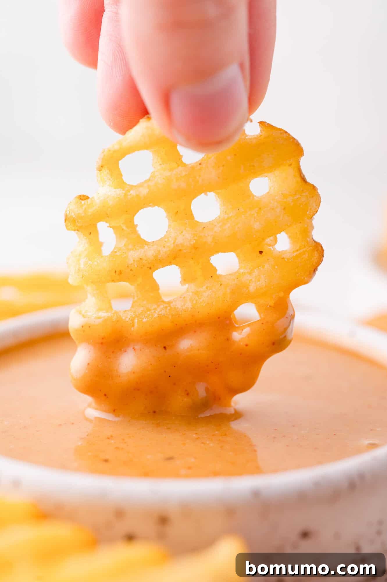 A hand dips a waffle-cut French fry into a bowl of creamy, orange-colored Chick-fil-A dipping sauce.
