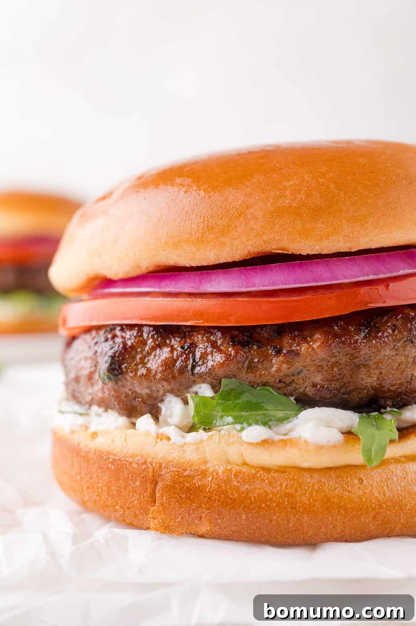 A close-up of a perfectly grilled lamb burger featuring vibrant sliced red onion, ripe tomato, crisp lettuce, and a generous dollop of creamy sauce, all nestled within a glossy brioche bun on white parchment paper.