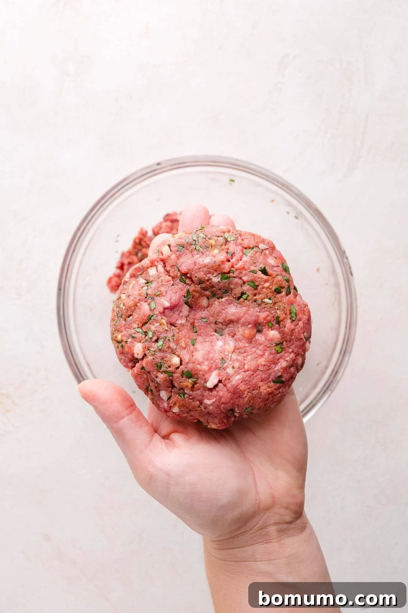 A skilled hand gently holds a perfectly shaped, raw, and seasoned ground lamb patty, poised just above a clear glass bowl containing the remaining mixture. The scene is set on a light-colored surface, highlighting the patty's readiness for cooking.