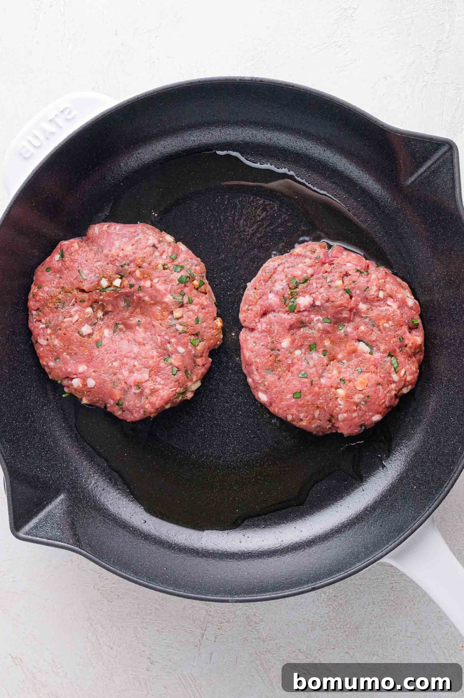 Two raw seasoned burger patties are cooking in a black skillet with a small amount of oil.