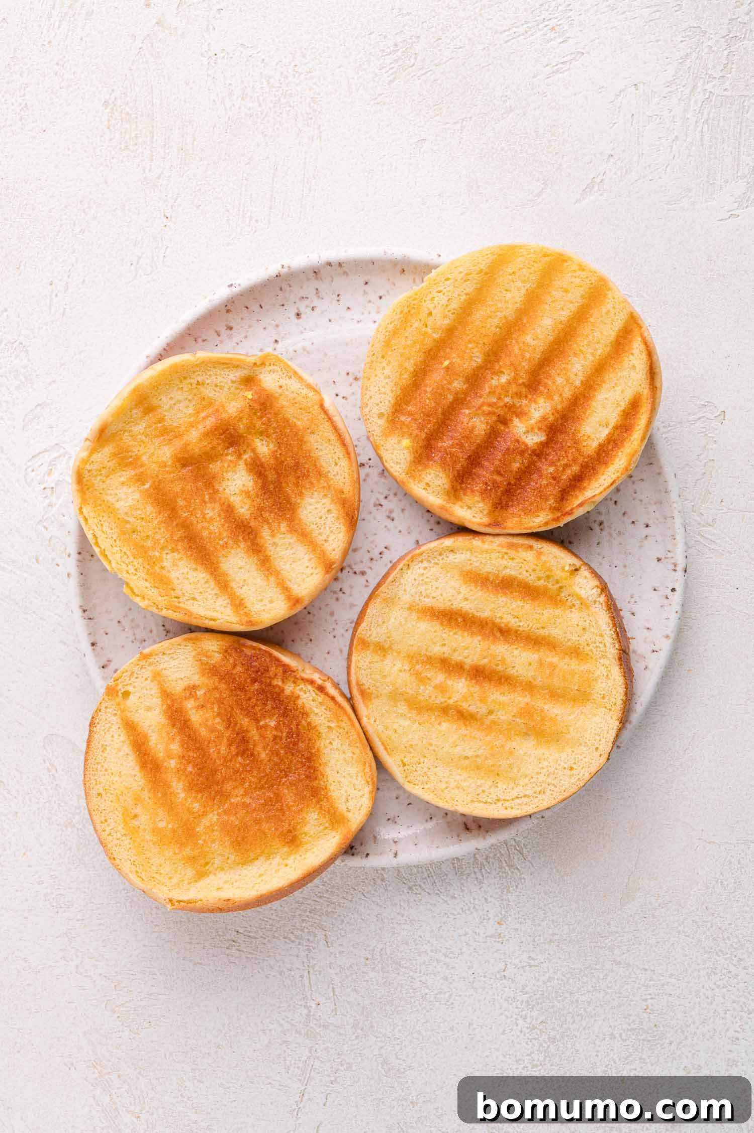 A plate with four toasted burger bun halves, showing grill marks on a light textured surface.