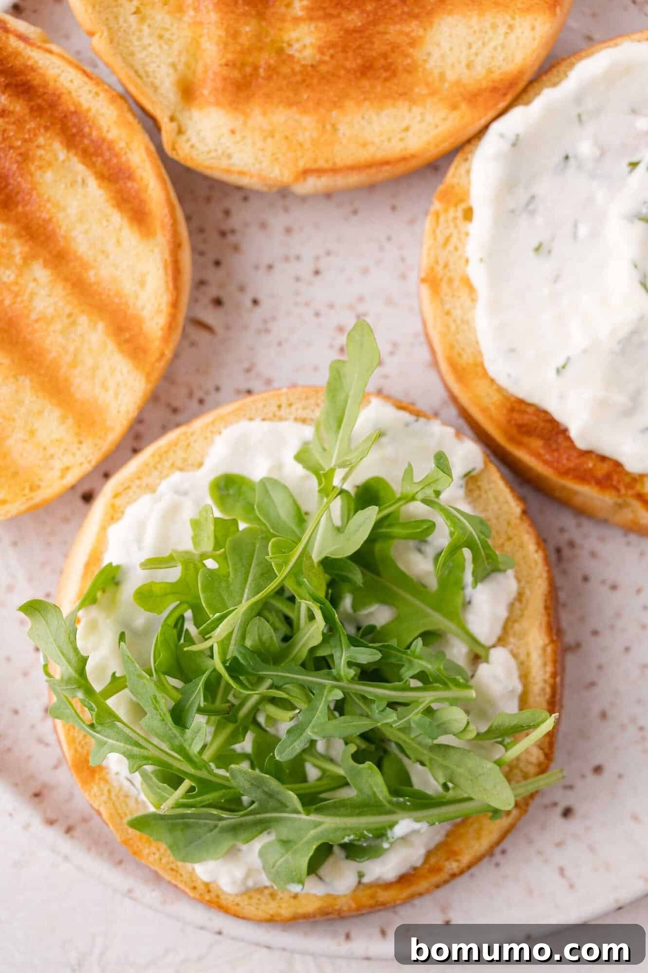 A toasted sandwich bun topped with a white spread and fresh arugula, next to other toasted buns with spread, on a speckled plate.