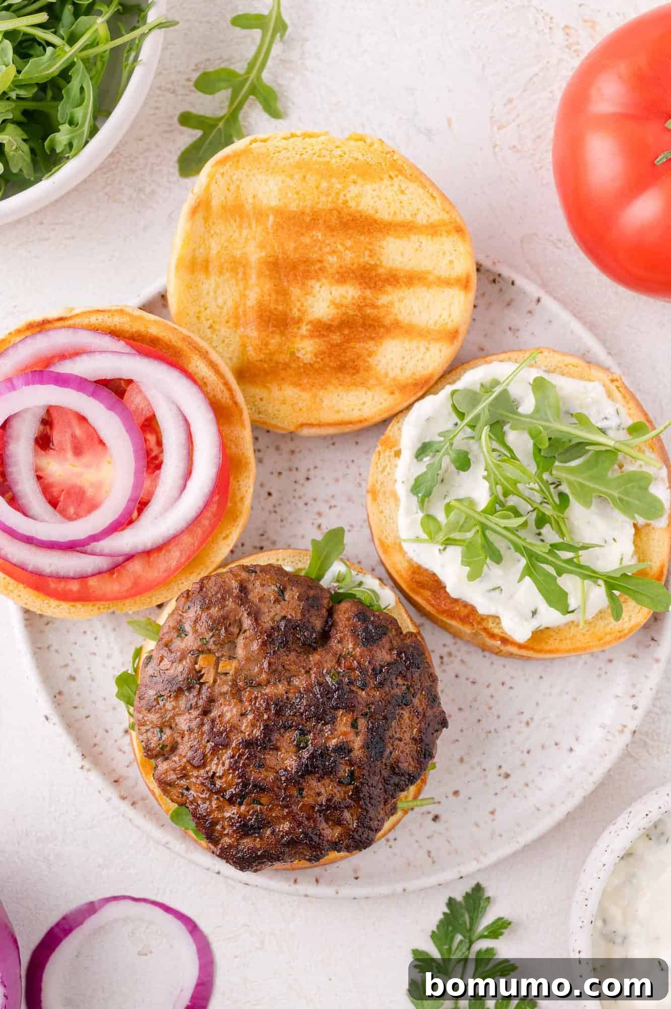 Open-faced burger on a plate with a grilled patty, arugula, sauce, tomato slices, and red onion on a toasted bun. Nearby are arugula, tomato, and onion slices.