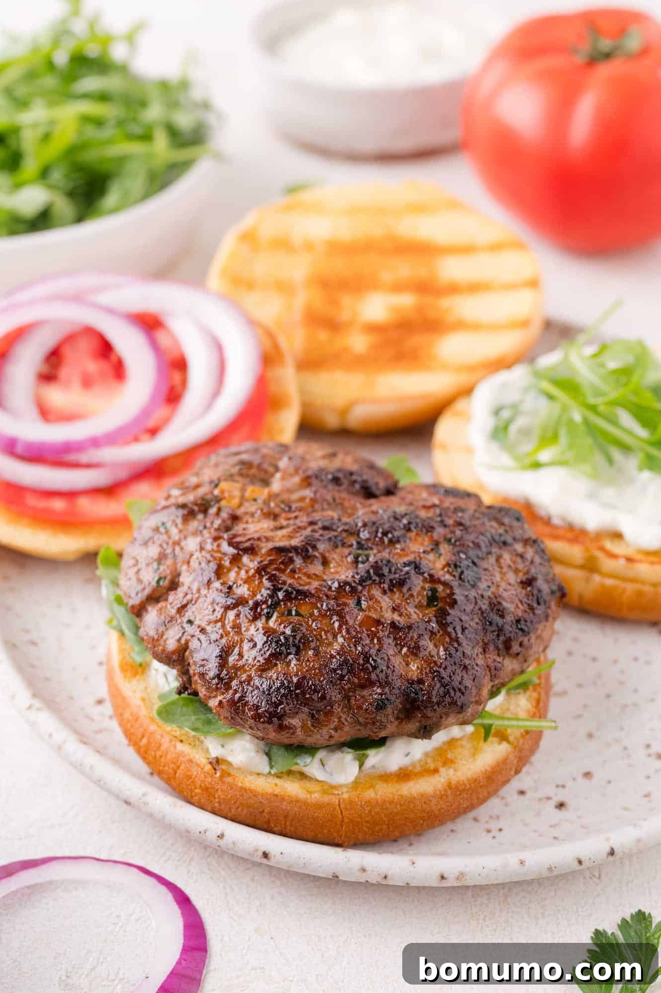 A grilled lamb burger patty artfully arranged on a bun with crisp greens and sauce, alongside perfectly sliced tomatoes, vibrant red onion, and another bun half with sauce and greens, all resting on a rustic plate with fresh, complementary ingredients nearby.
