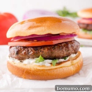 A perfectly cooked lamb burger with sliced tomato, red onion, lettuce, and creamy white sauce, all neatly assembled in a toasted bun and presented on a clean white surface.