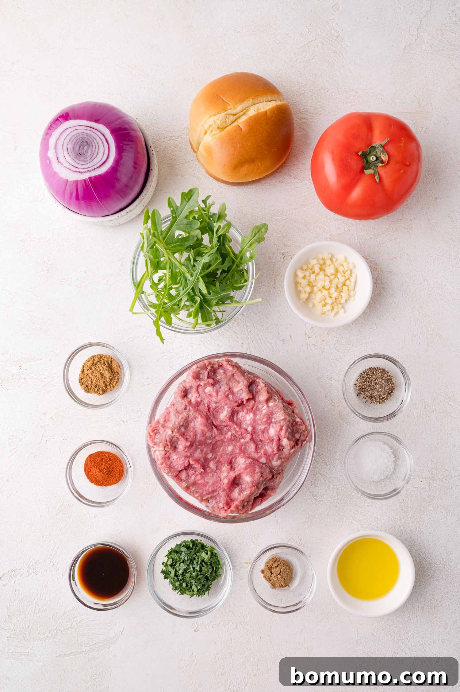 A vibrant collection of fresh ingredients for making delicious lamb burgers, meticulously arranged on a clean white surface. Components include rich ground lamb, a soft burger bun, plump tomato slices, thinly sliced red onion, fresh arugula, aromatic minced garlic, a selection of vibrant spices, fresh herbs, a bottle of Worcestershire sauce, golden olive oil, and essential salt and pepper.