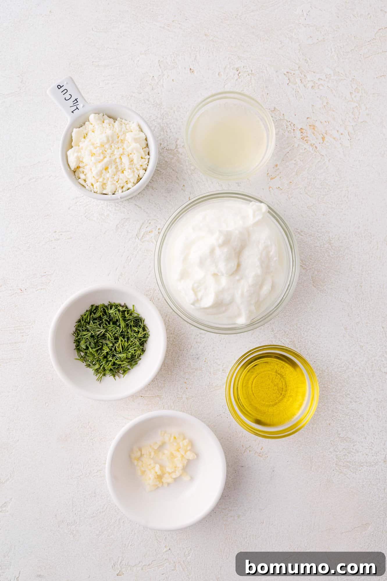Five small, elegant bowls contain individual ingredients for the sauce: crumbled feta cheese, fresh lemon juice, creamy Greek yogurt, finely chopped herbs, rich olive oil, and pungent minced garlic. They are neatly arranged on a light-textured surface, ready for mixing.